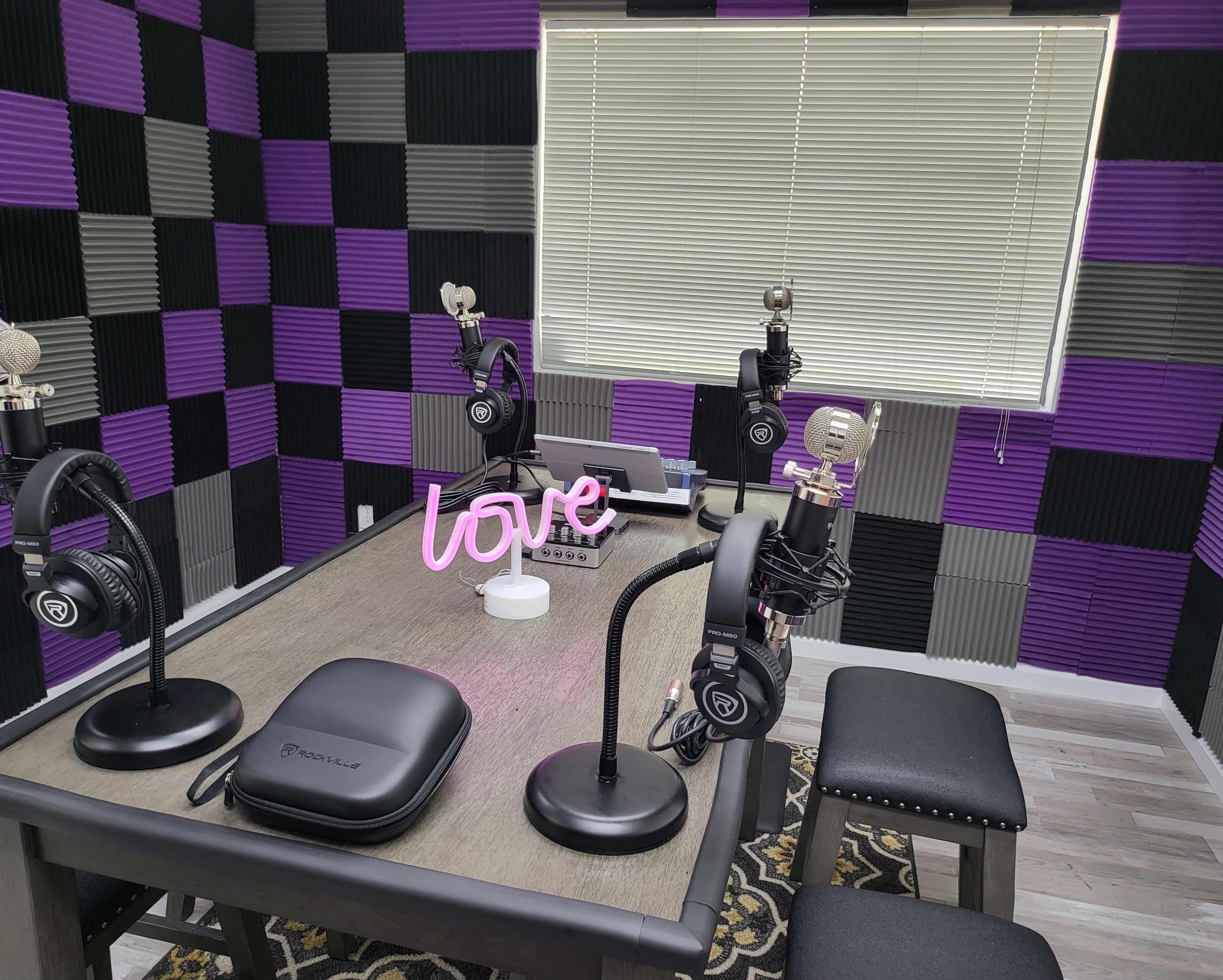 A podcast studio setup with four microphones, headphones, and a decorative "love" sign on a table, surrounded by purple and gray soundproofing panels.