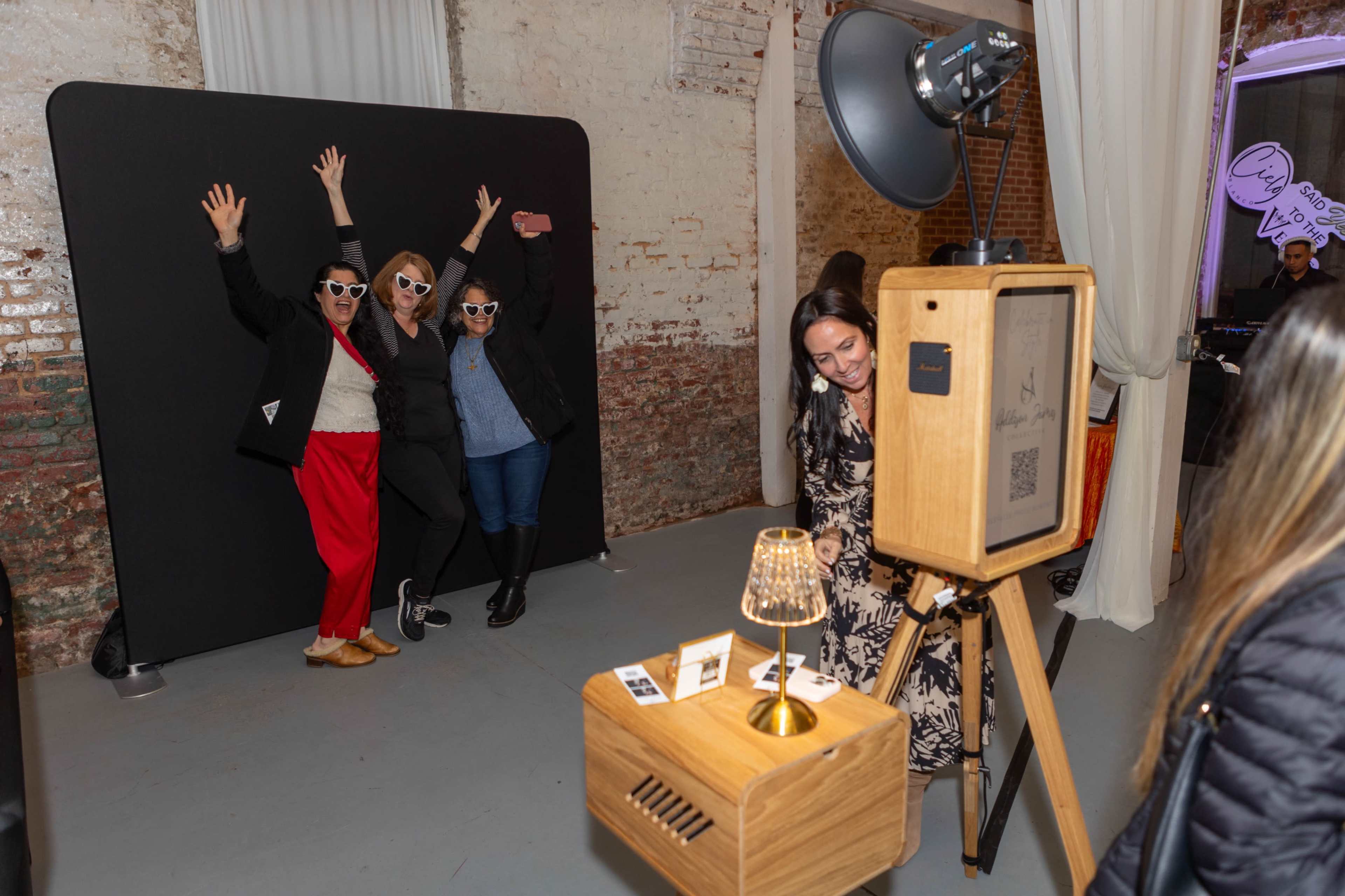 Three people are posing with their hands raised in front of a photo booth backdrop while another person is interacting with a photo printing station nearby.