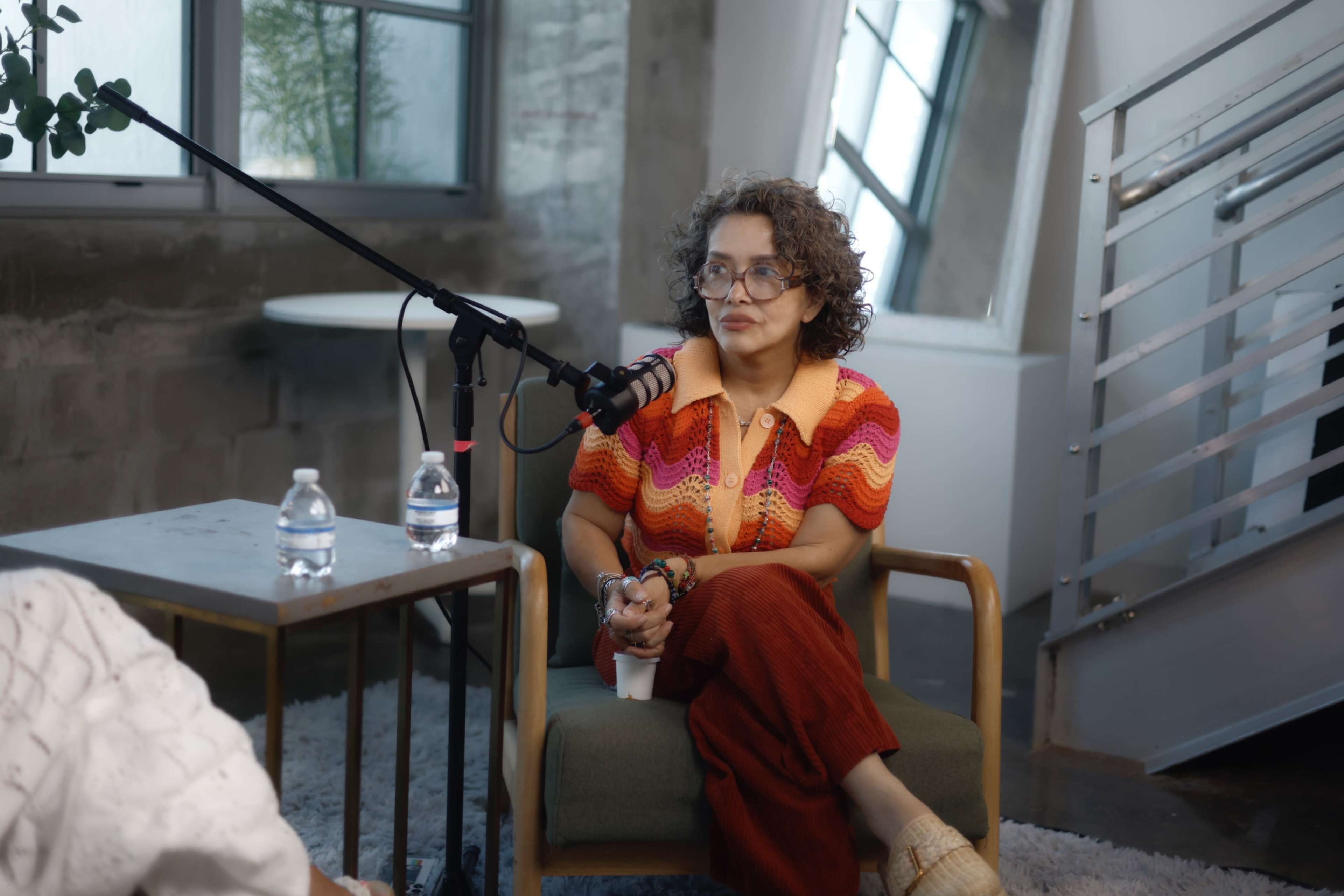 A person with curly hair is sitting on a chair in a well-lit room, engaged in an interview with a microphone positioned nearby and bottles of water on a table.