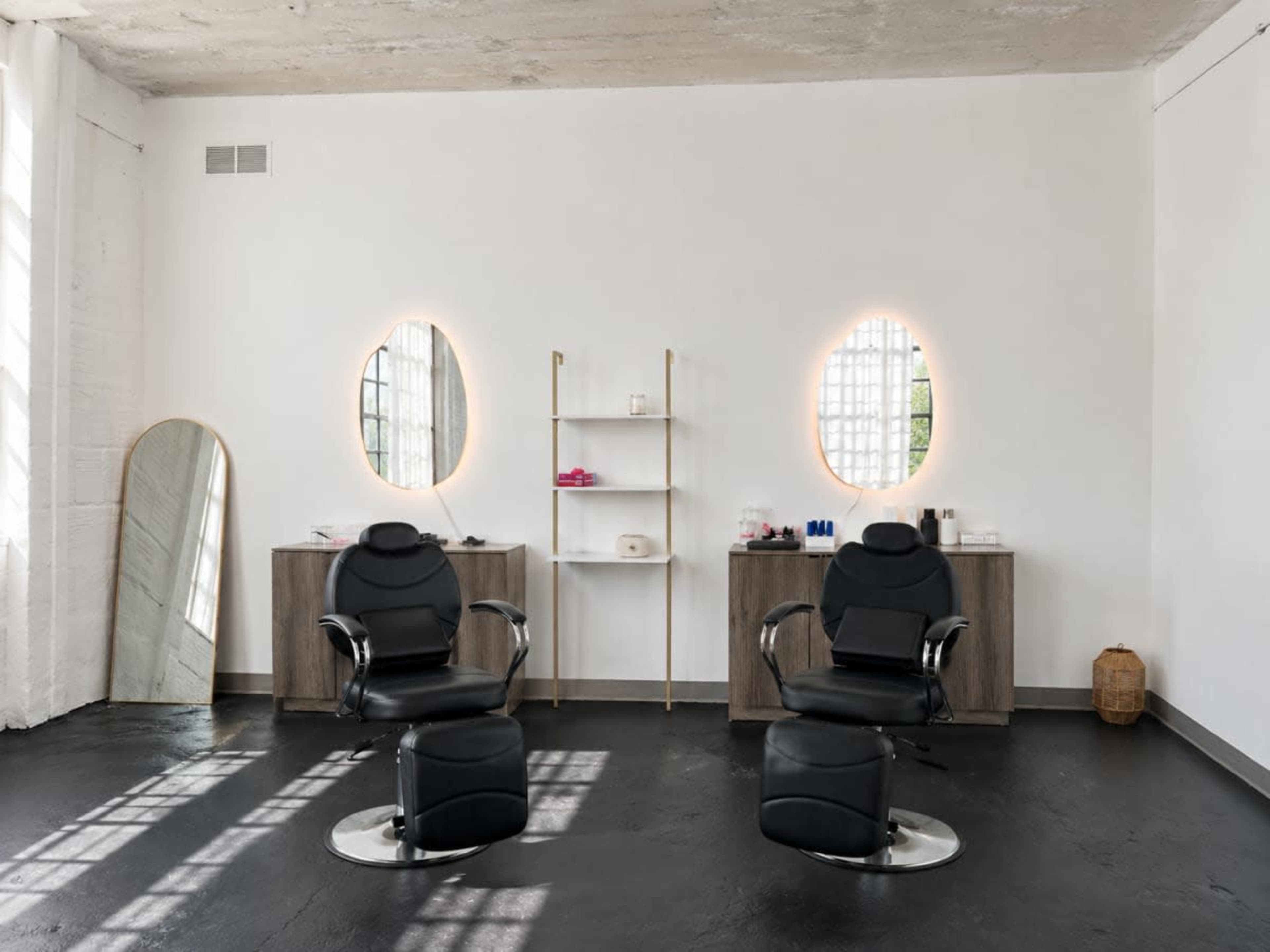 The image shows a minimalist hair salon with two black styling chairs and a small shelf against a white wall, featuring oval mirrors and natural light streaming through windows.