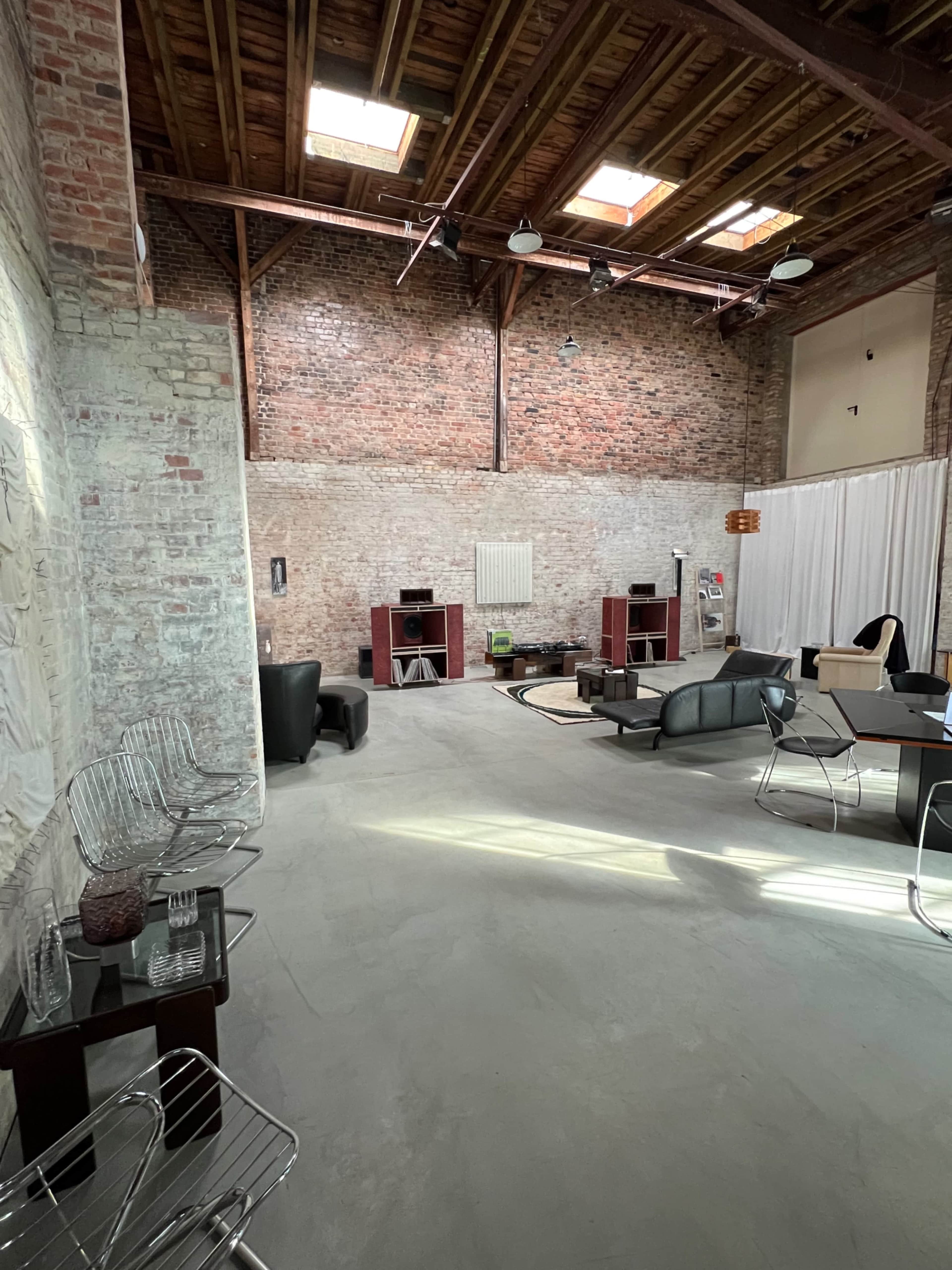 The image shows a spacious industrial-style living area with exposed brick walls, wooden beams, and modern furniture arranged around a central coffee table.