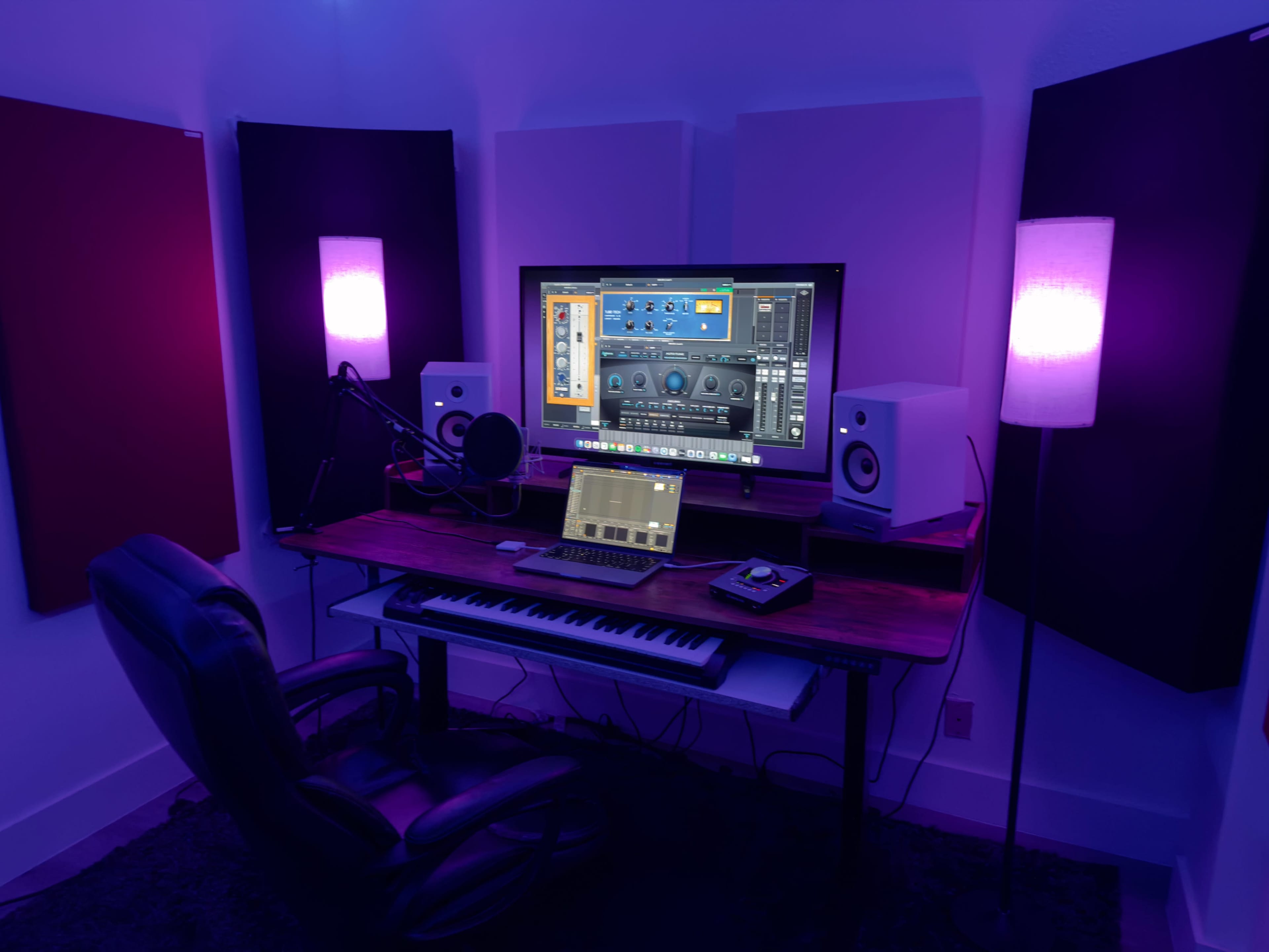 The image shows a music production studio with a desk featuring a computer monitor, audio interface, keyboard, and studio monitors, surrounded by sound-absorbing panels.