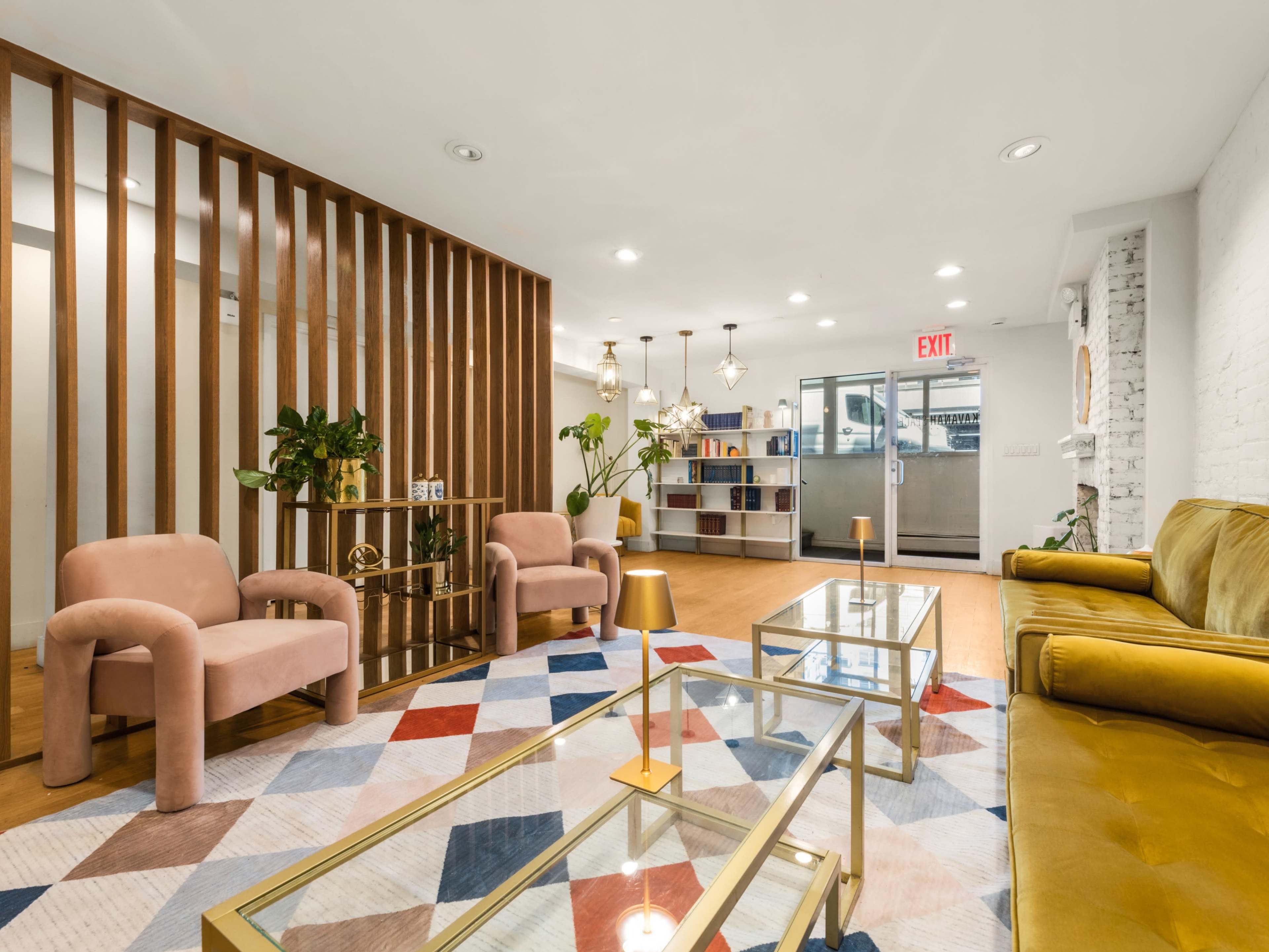 The image shows a modern lounge area featuring two pink armchairs, a yellow couch, a geometric-patterned rug, and a bookshelf against the wall.