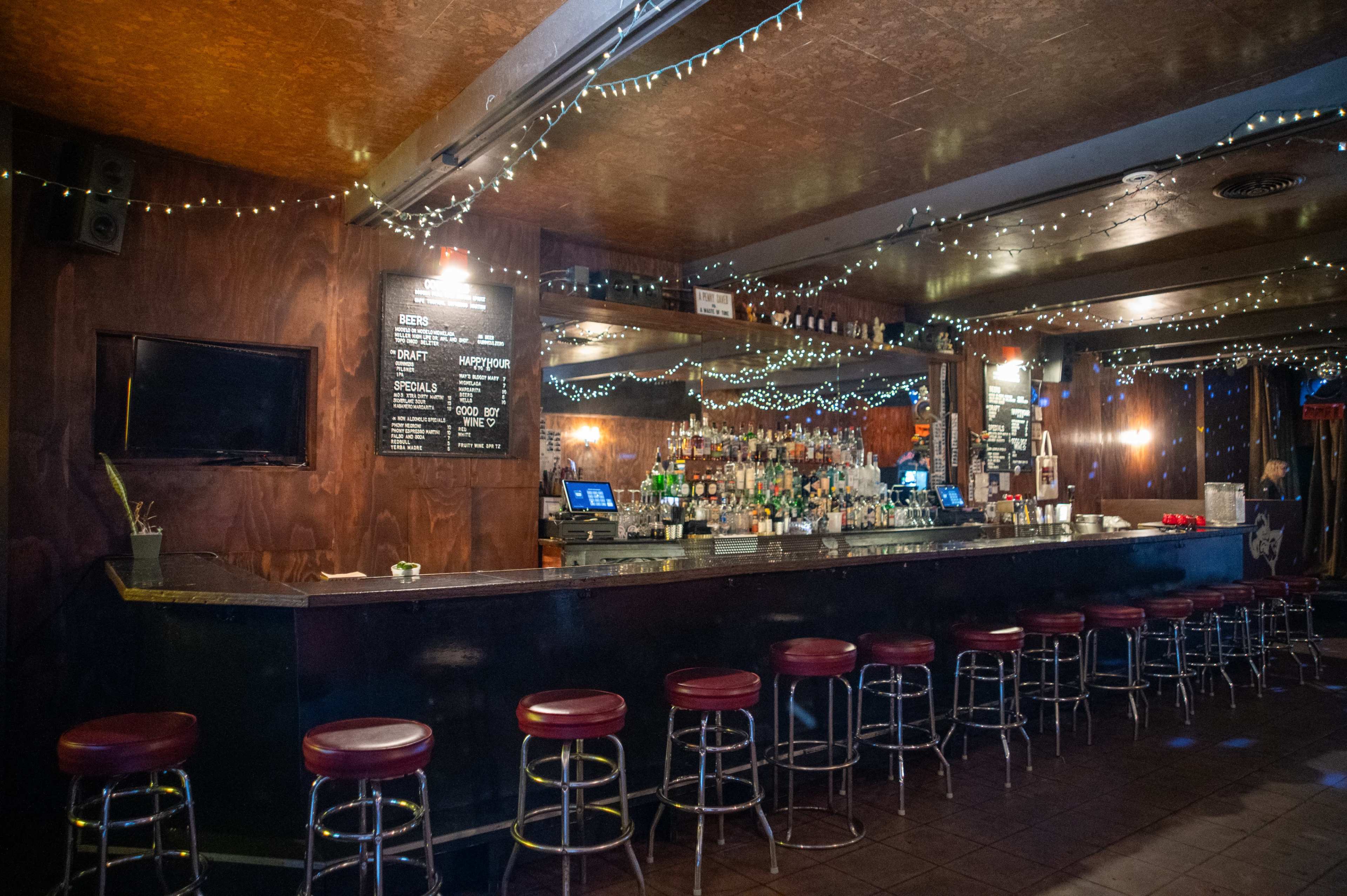 The image shows a dimly lit bar with a long, dark countertop, red stools, and decorative string lights overhead.