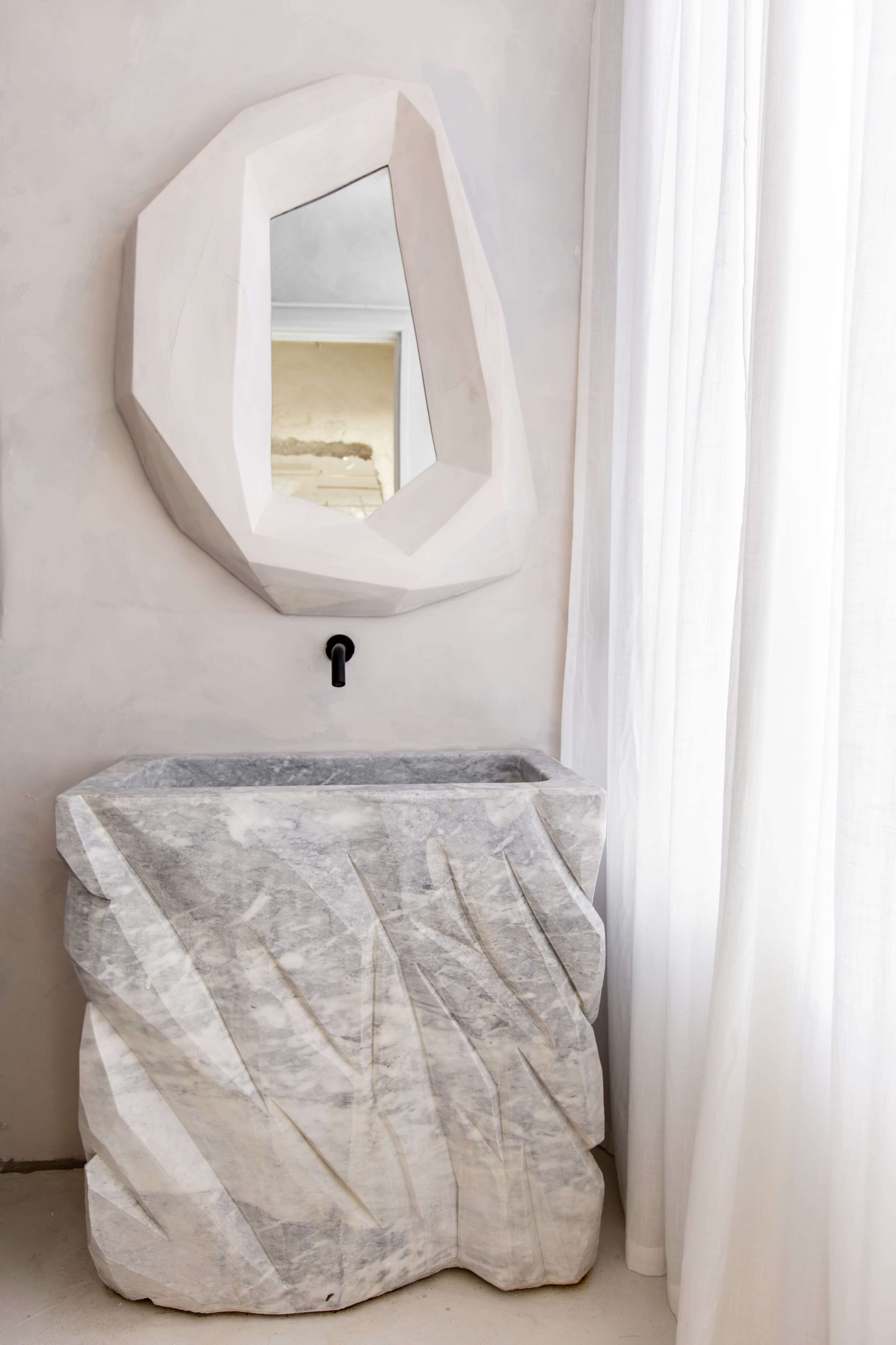 The image features a sculptural marble sink with a textured finish and a geometric mirror mounted above it, set against a pale wall and soft white curtains.