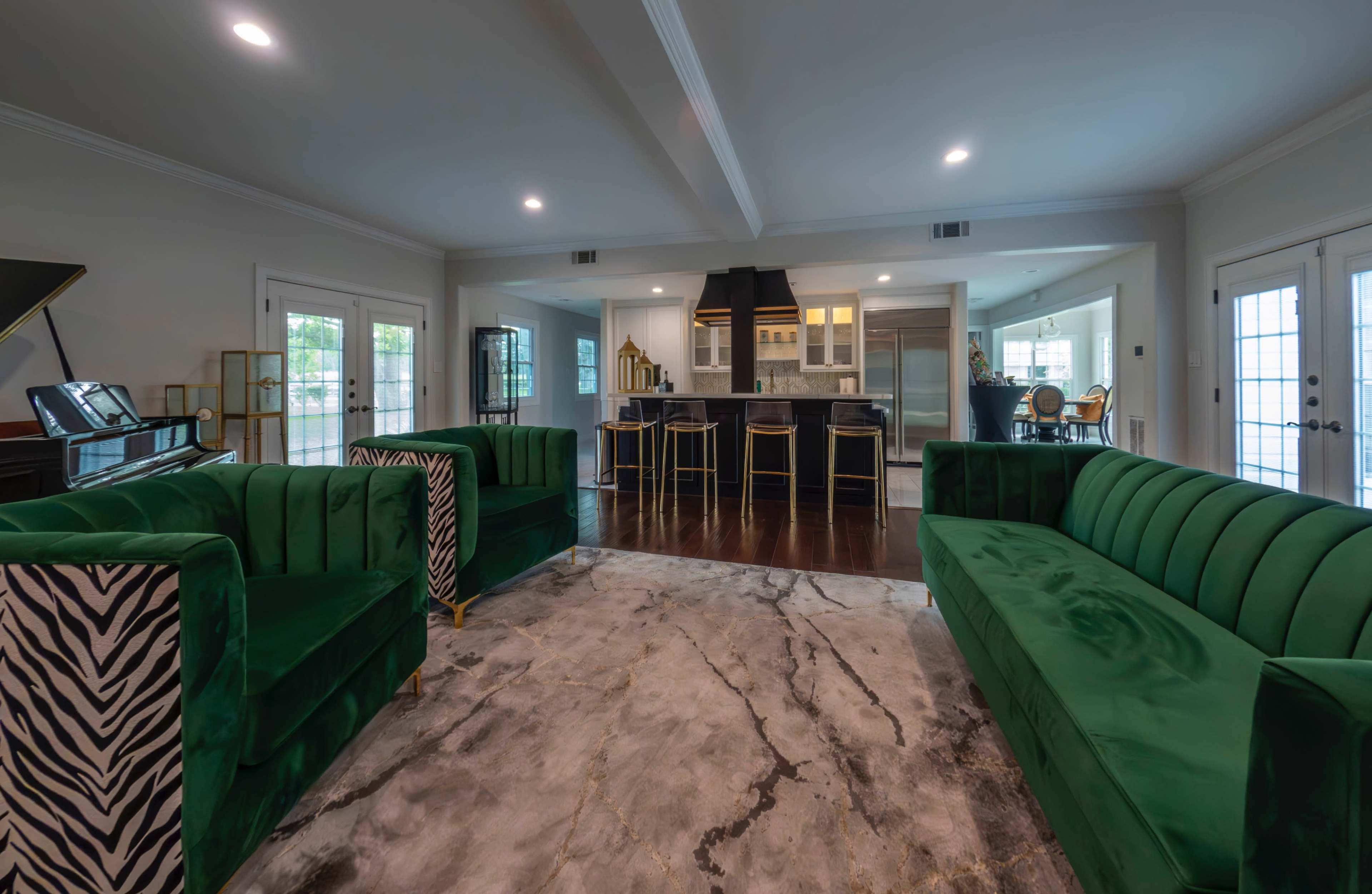 The image shows a spacious living area with two green velvet sofas, a marble rug, and a kitchen with dark cabinetry in the background.