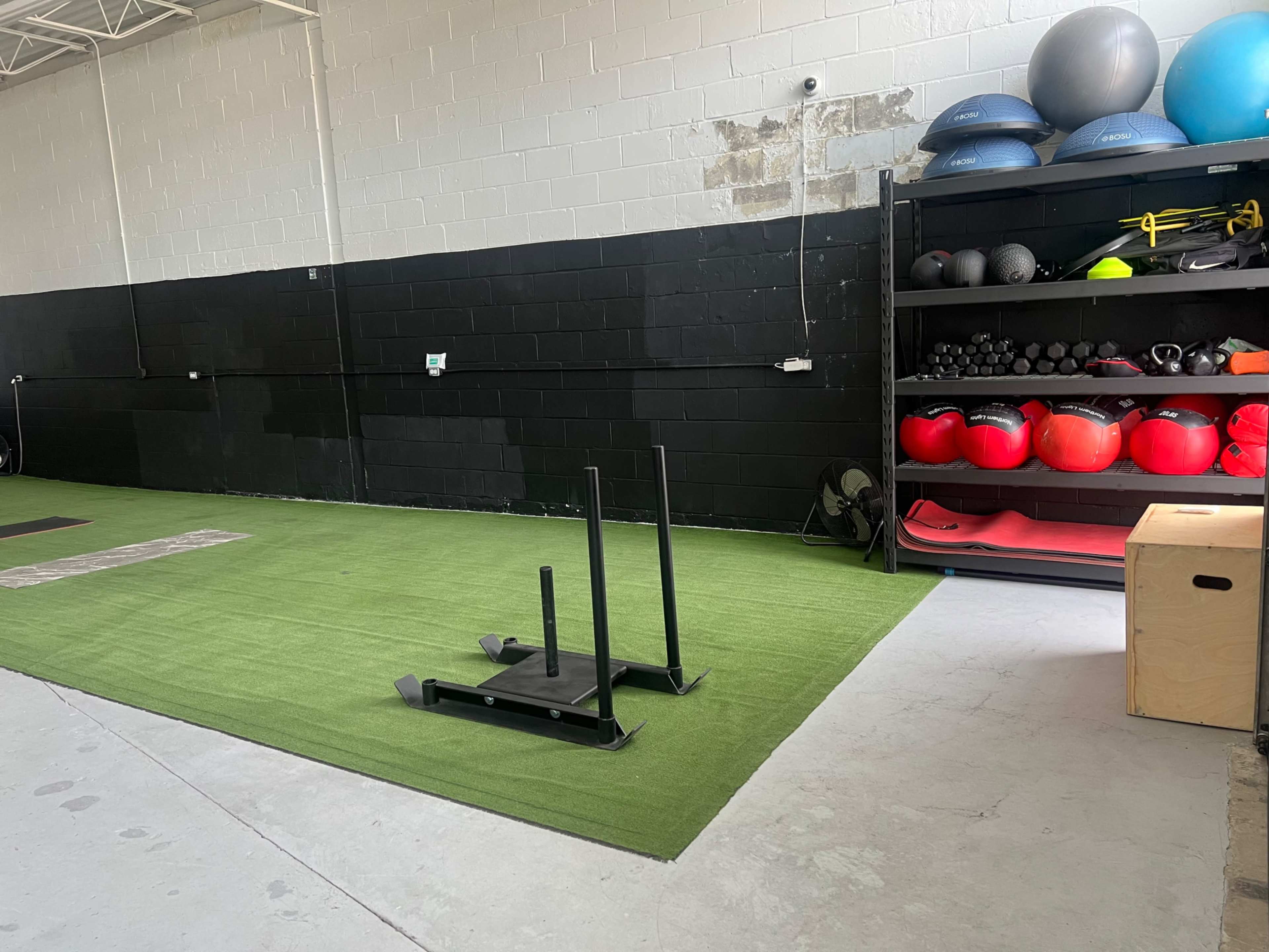 The image shows a gym area with a green turf surface, a sled for pushing, a storage shelf with various exercise equipment, and a wooden box in the corner.
