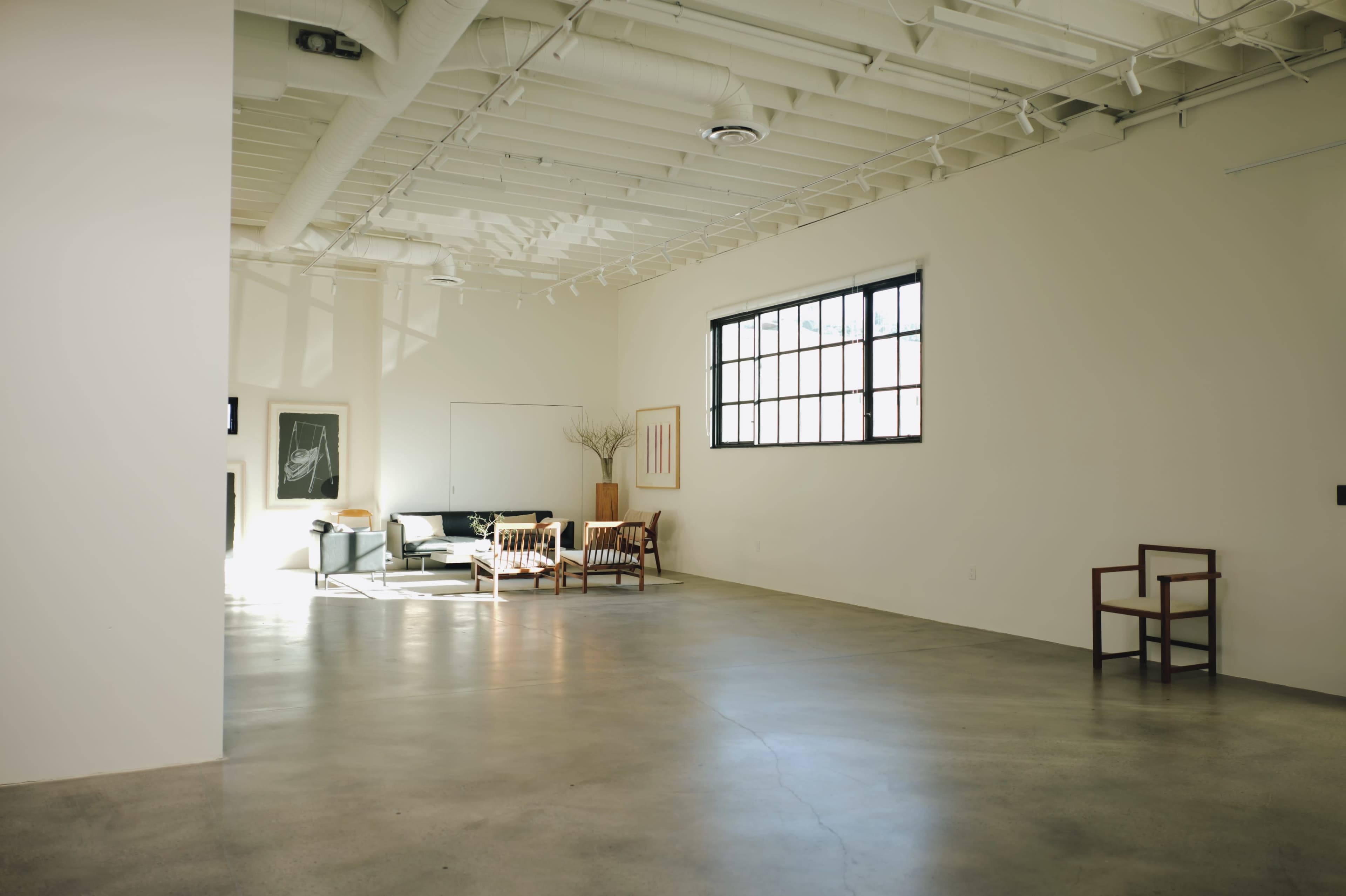 The image shows a spacious, minimalist room with polished concrete flooring, a large window, and a few pieces of modern furniture including a sofa and chairs.