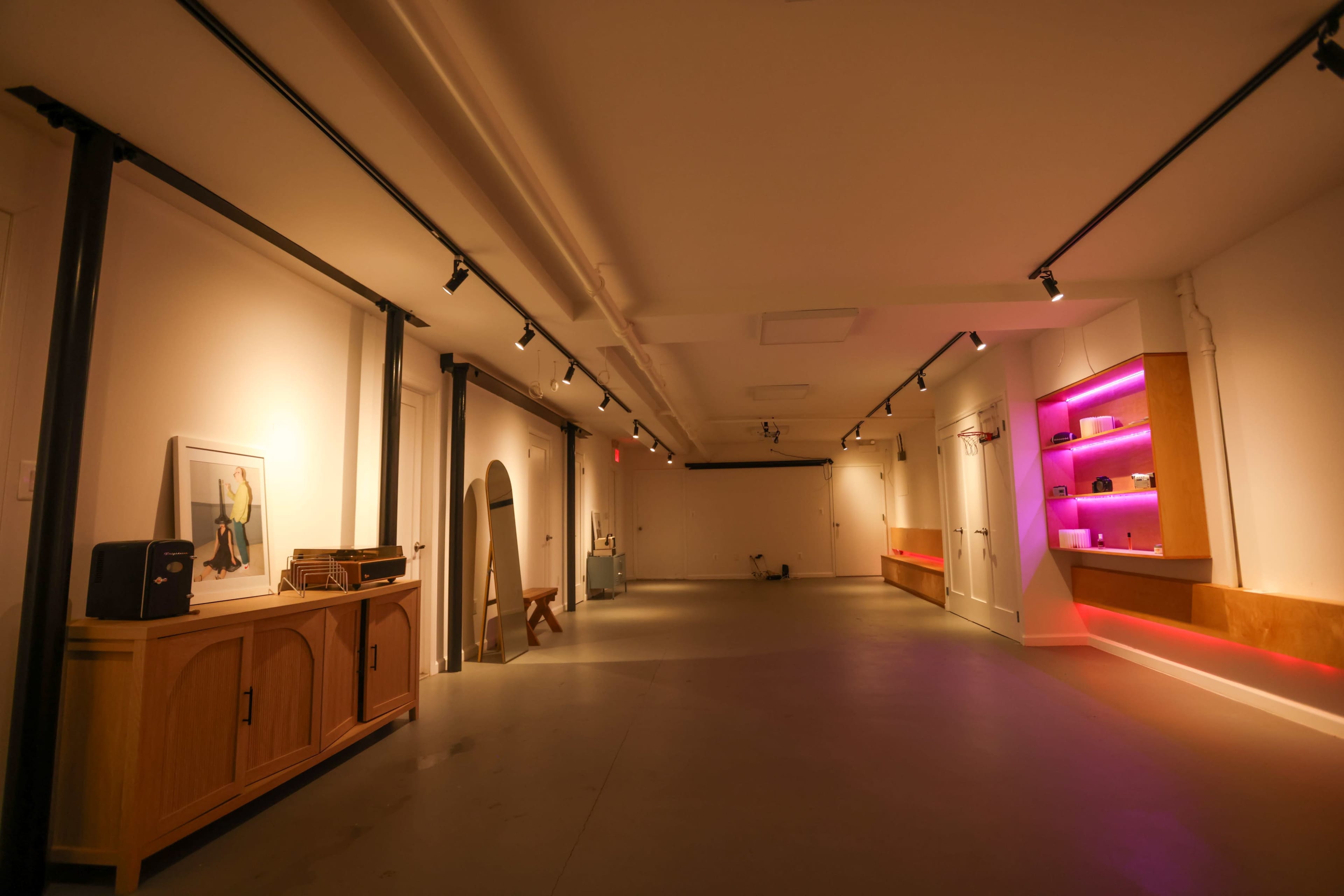 The image shows a spacious, well-lit interior with a minimalist design, featuring wooden furniture, a shelf with purple lighting, and smooth concrete flooring.