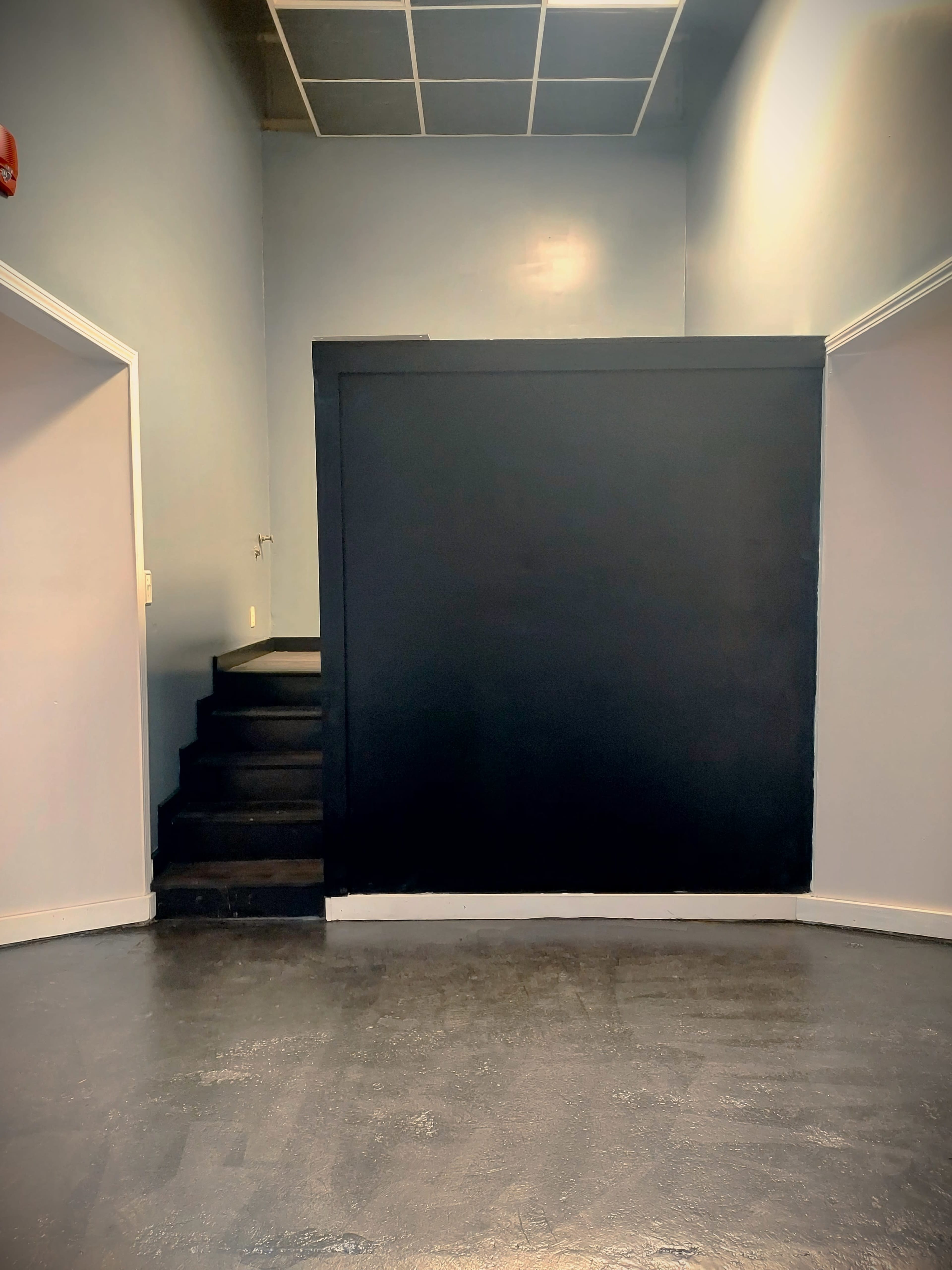 The image shows a dimly lit room with a black partition, a staircase on the left, and light-gray walls.