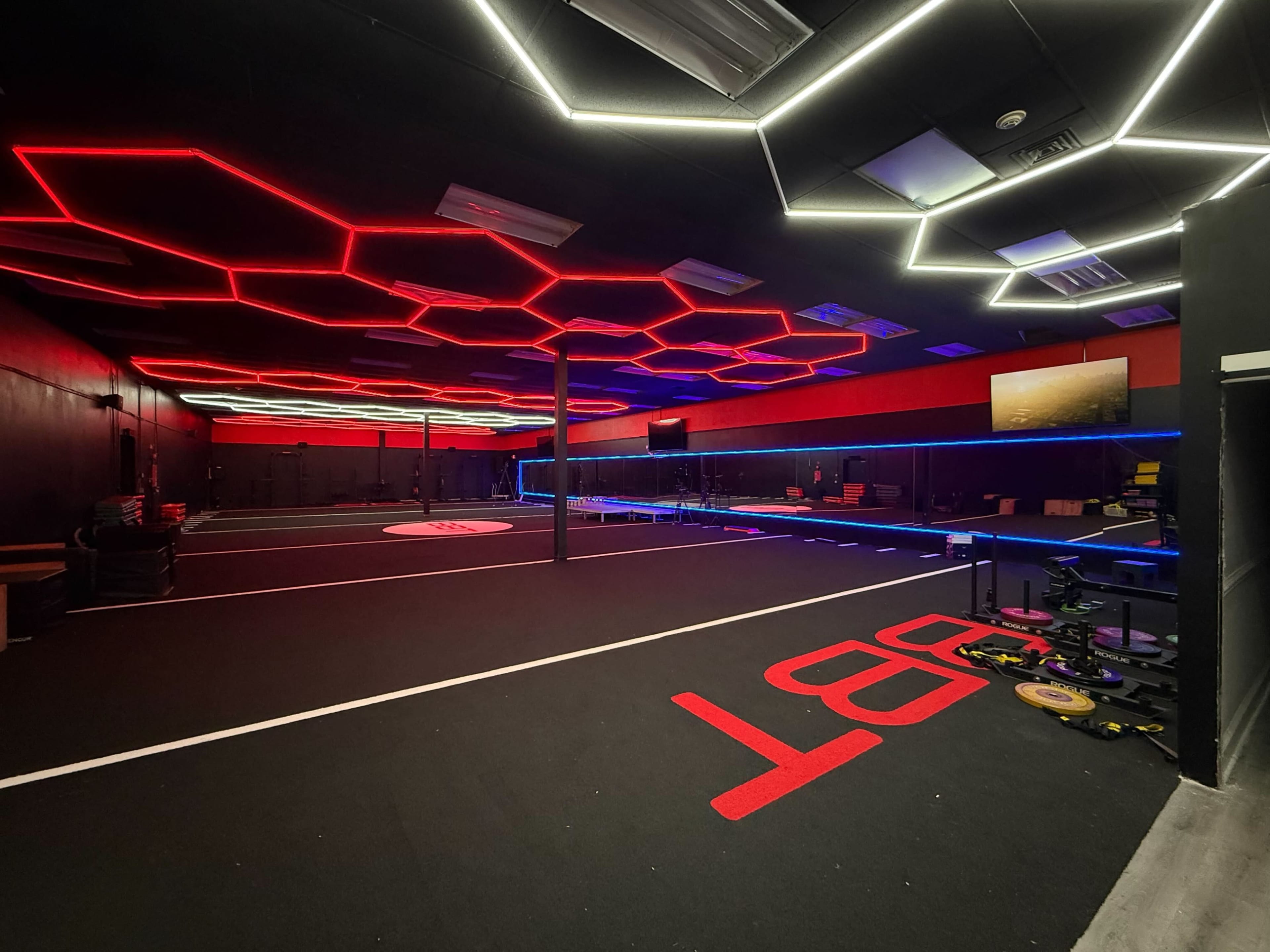 The image shows a modern gym interior with black walls, colorful LED lights in geometric patterns, and a spacious workout area featuring exercise equipment and flooring marked with red lettering.