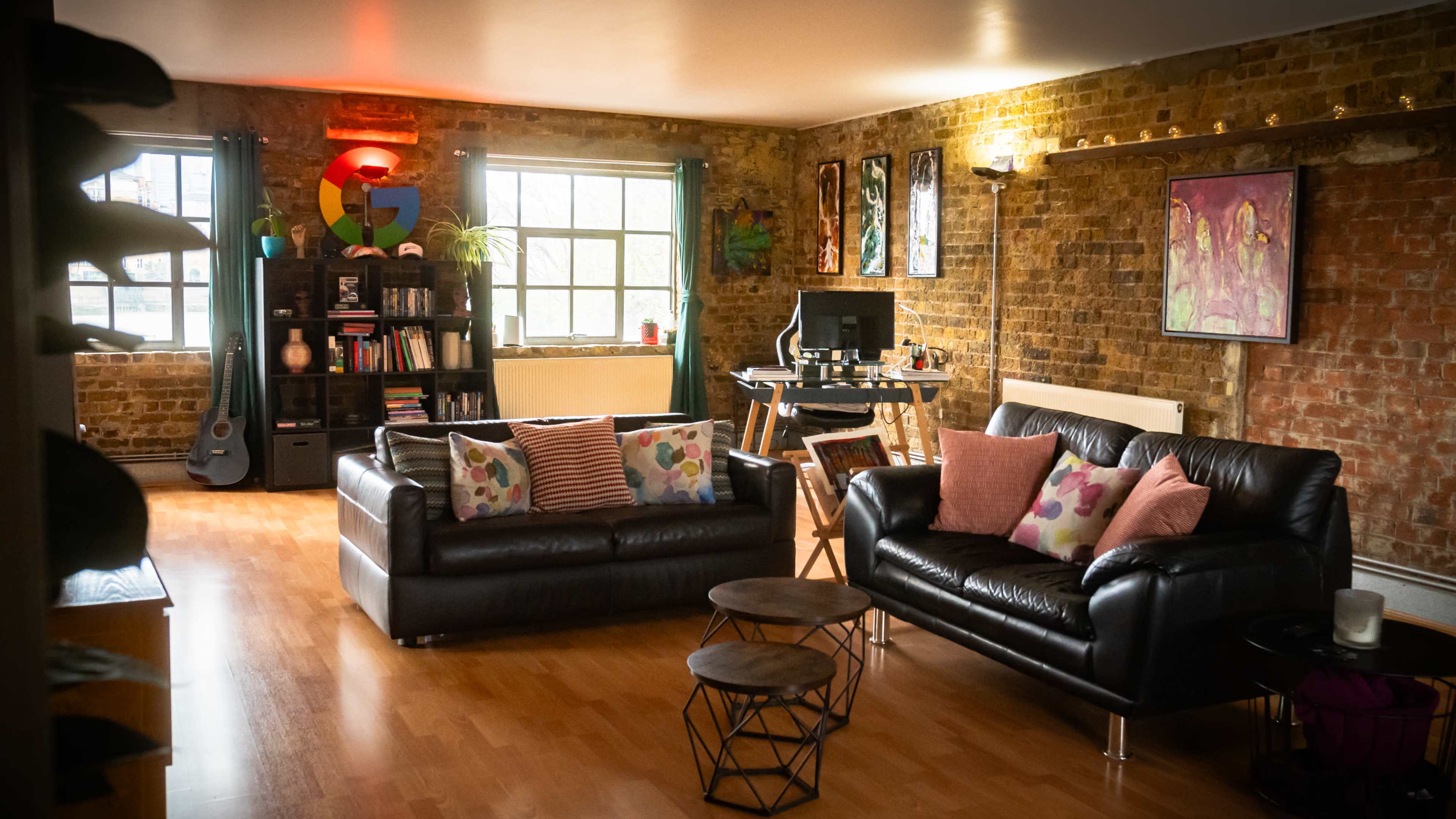 A cozy living room features two black sofas with colorful pillows, a wooden floor, a brick wall, and a small desk with a computer setup.