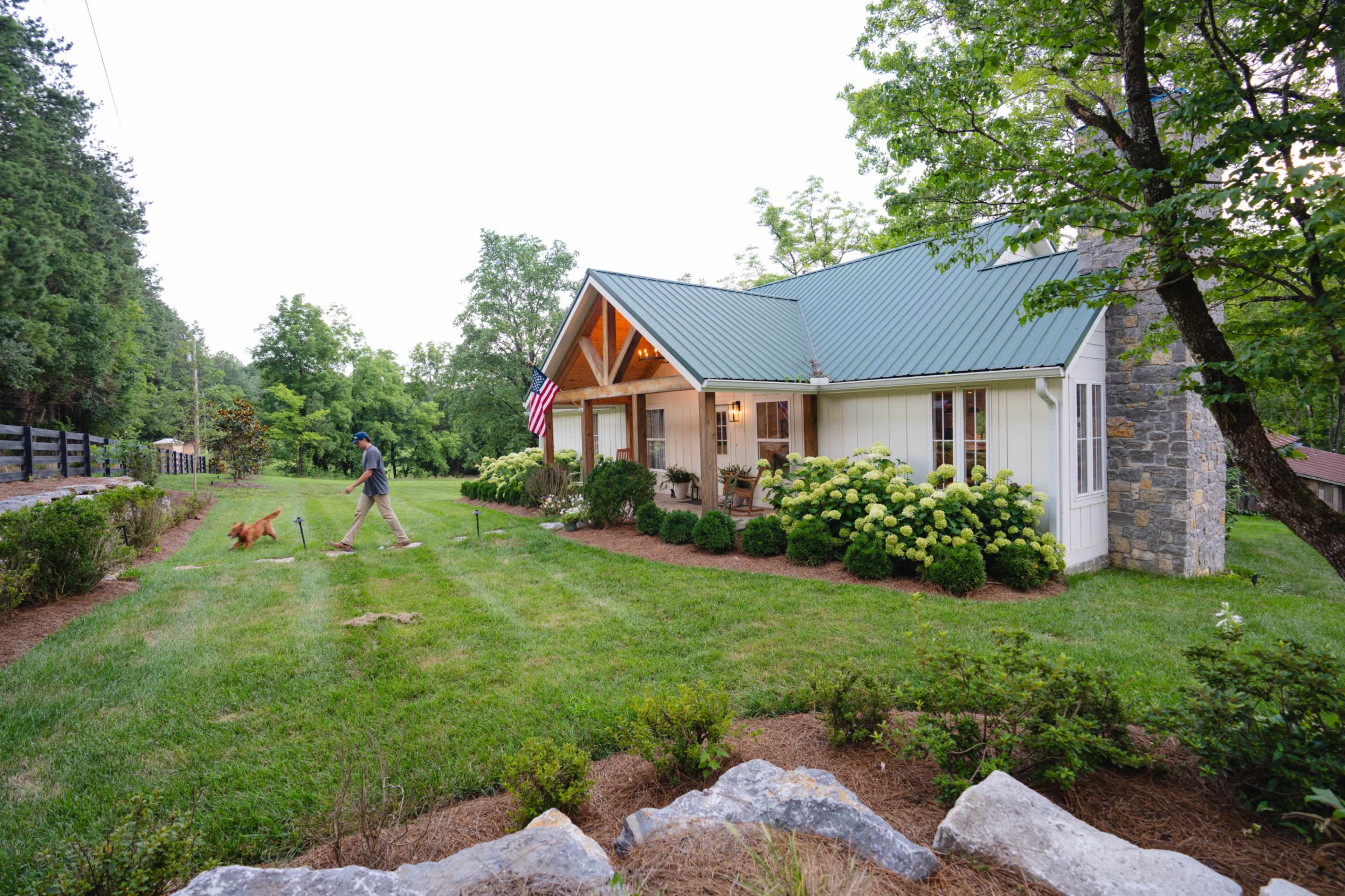 A person walks a dog on a lawn in front of a house with a stone chimney and a metal roof, surrounded by greenery and flowers.