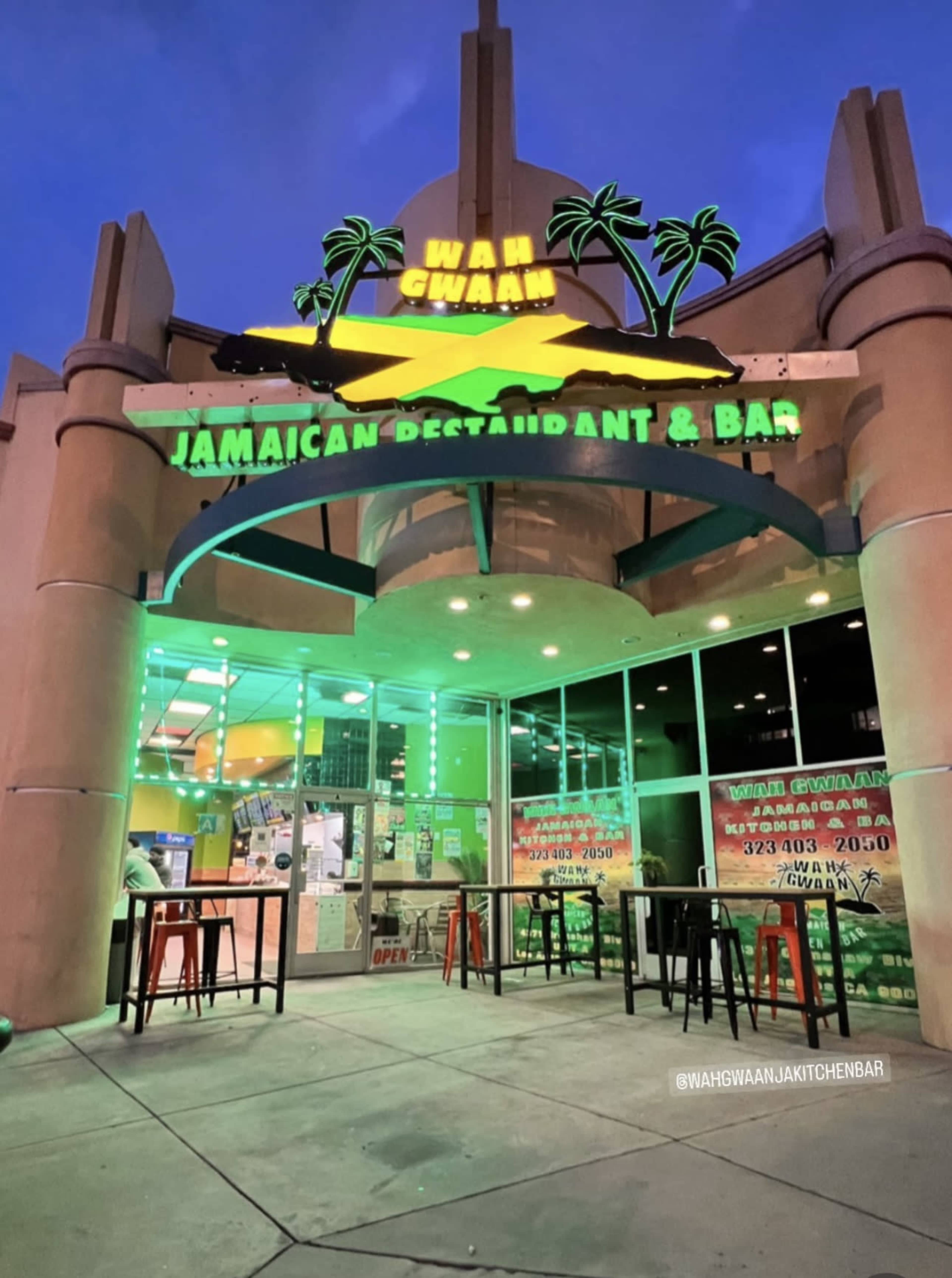 The image shows the vibrant exterior of a Jamaican restaurant and bar named "Wah Gwaan," featuring a neon sign and outdoor seating area.