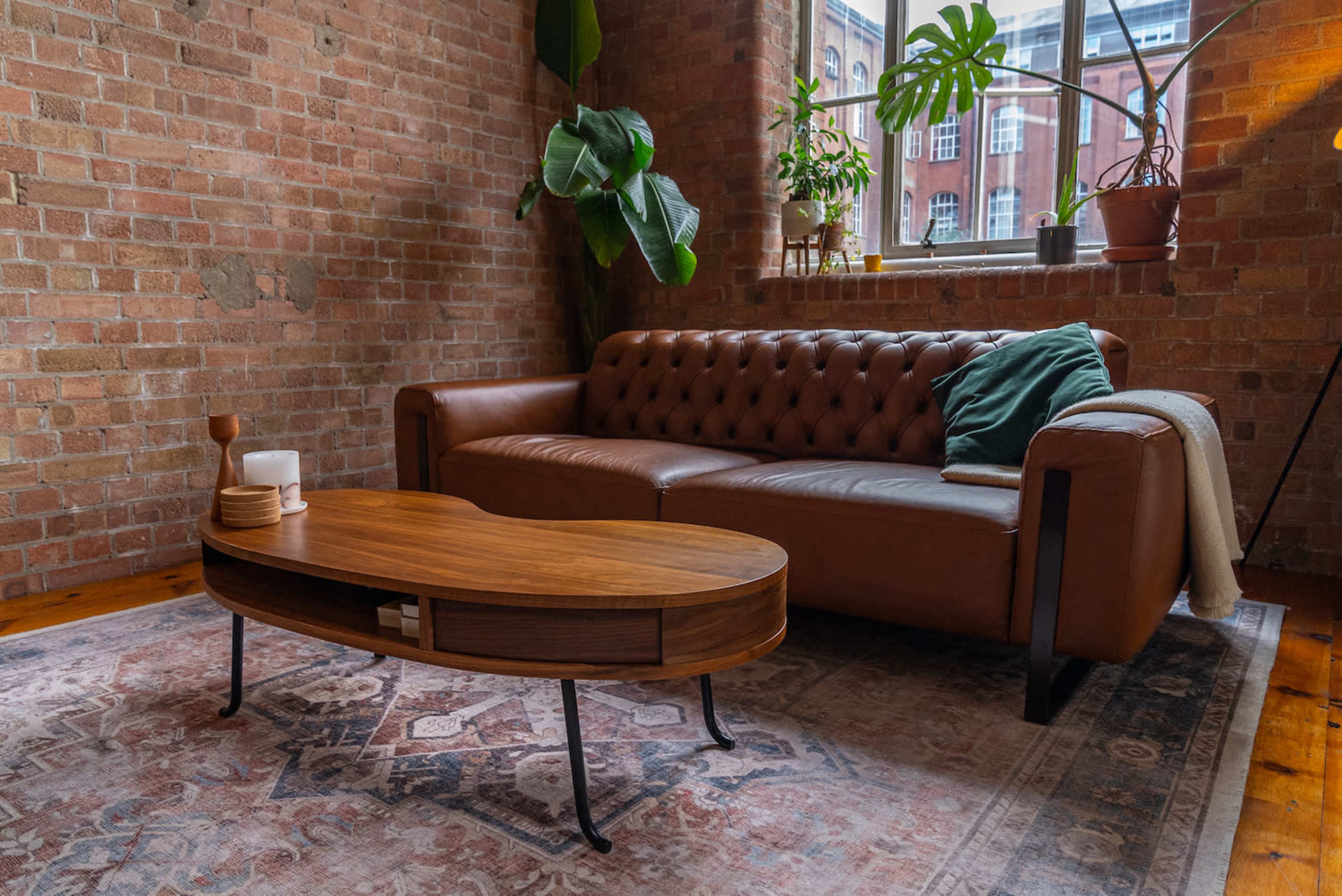 A brown leather sofa and a wooden coffee table in a room with exposed brick walls and a large window.