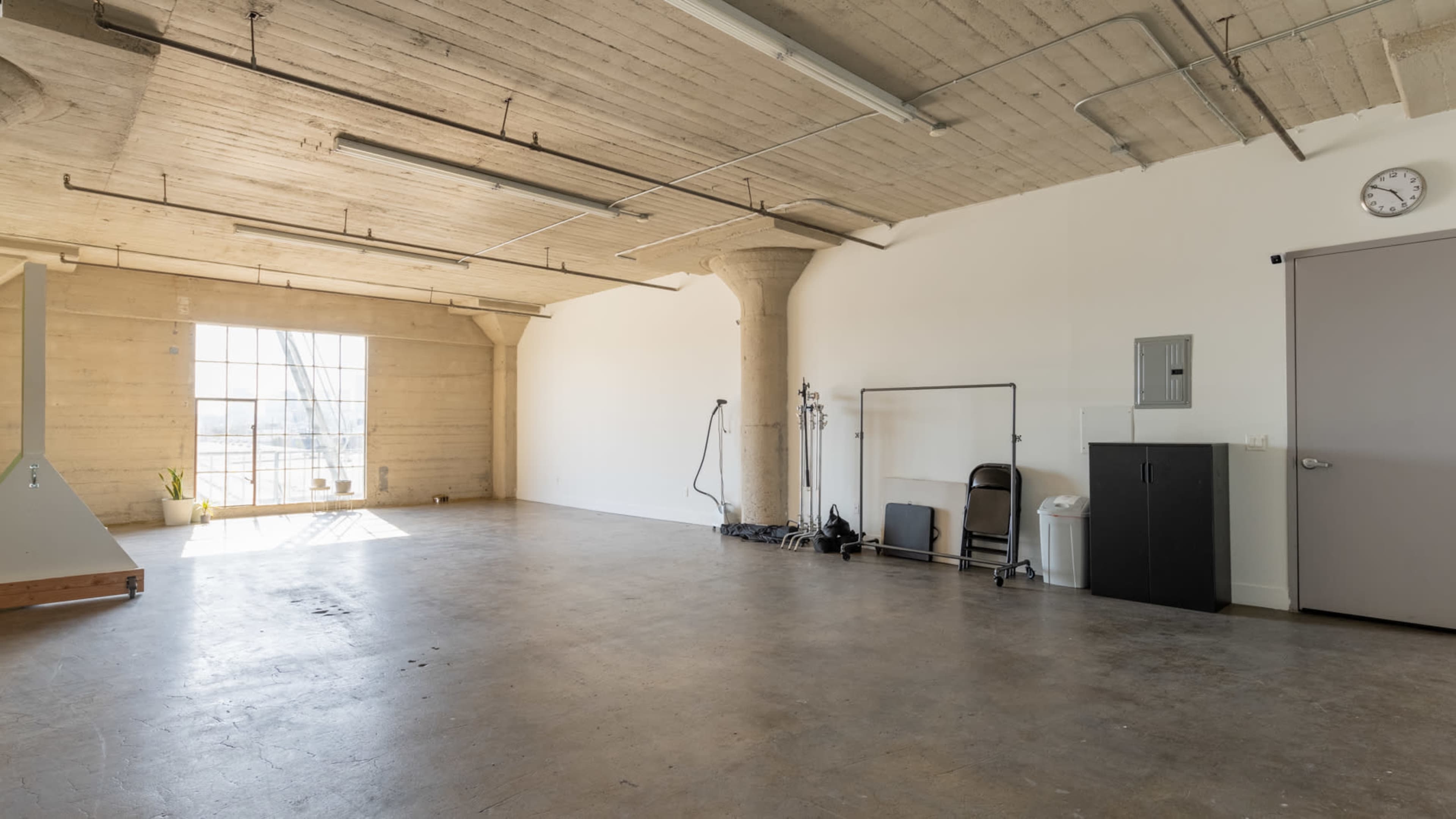 A spacious, empty room with concrete floors and large windows, featuring a few minimalist furnishings and industrial-style pillars.