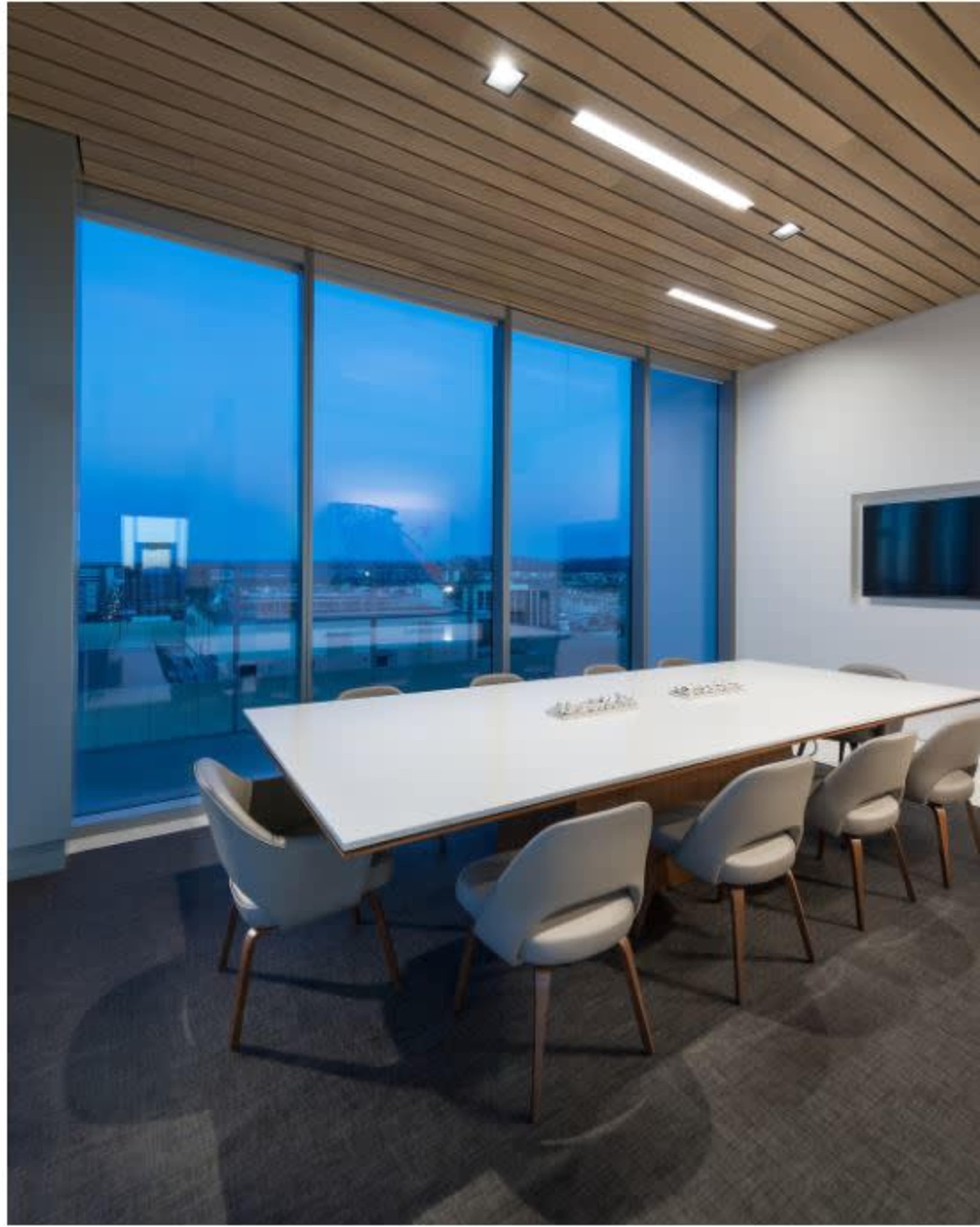 A modern conference room features a long white table surrounded by gray chairs, with large windows showcasing a blue evening sky.