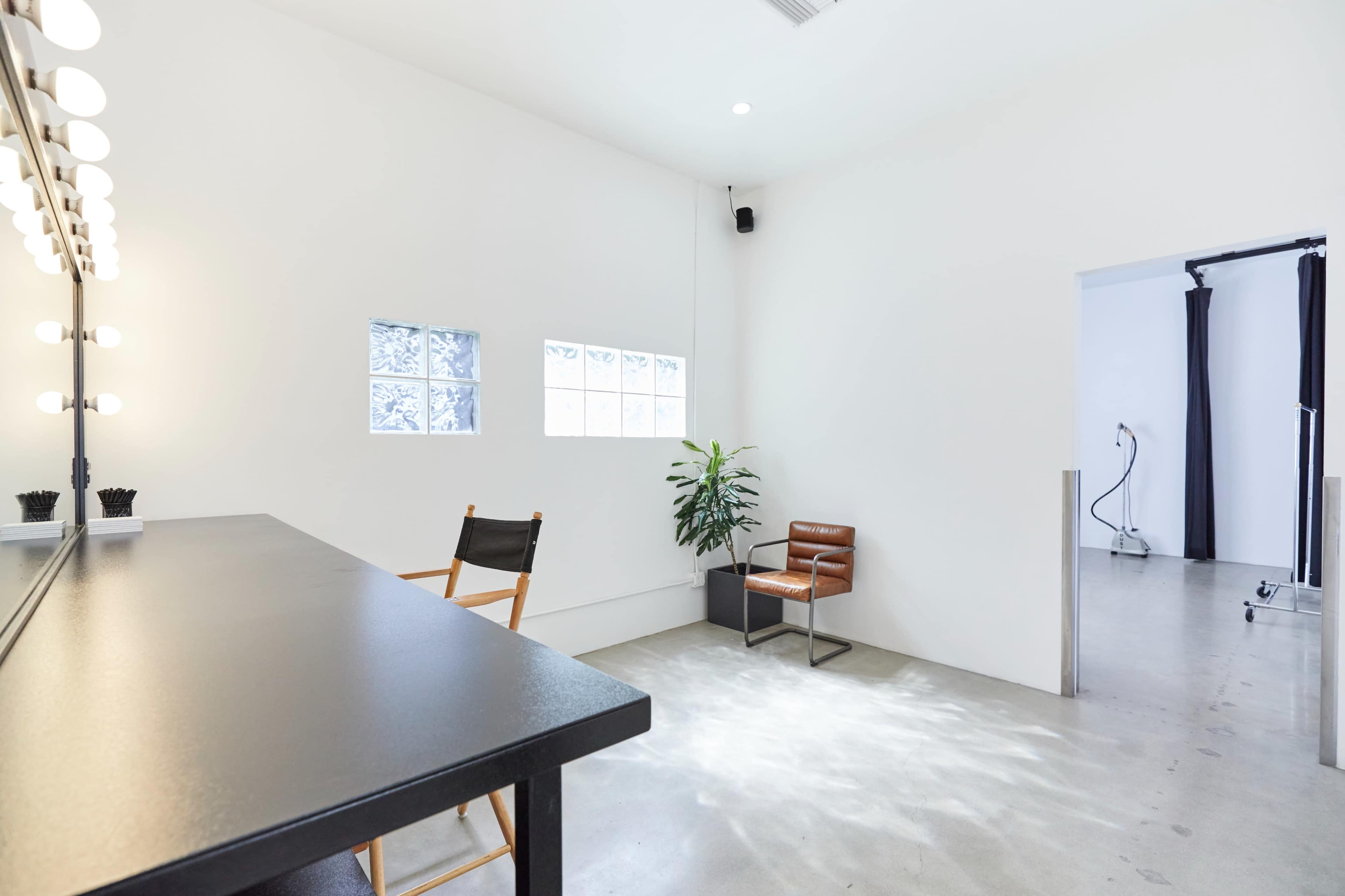 The image shows a minimalist dressing room with a large mirror illuminated by light bulbs, a single chair, a potted plant, and a doorway leading to another room.