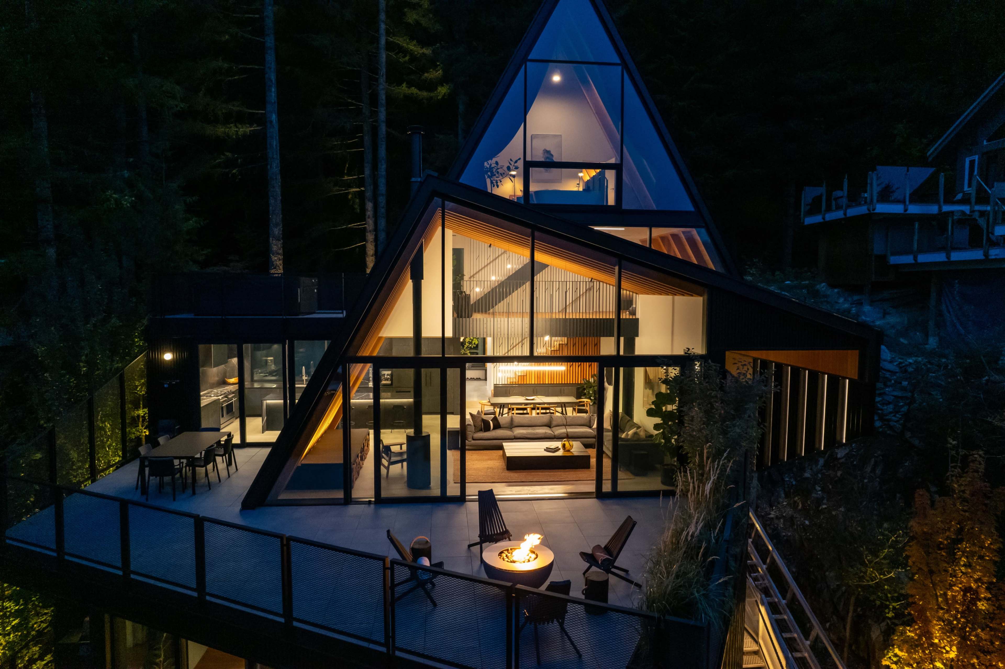 A modern A-frame house with large glass panels and a deck is illuminated by outdoor lighting at night, surrounded by trees.