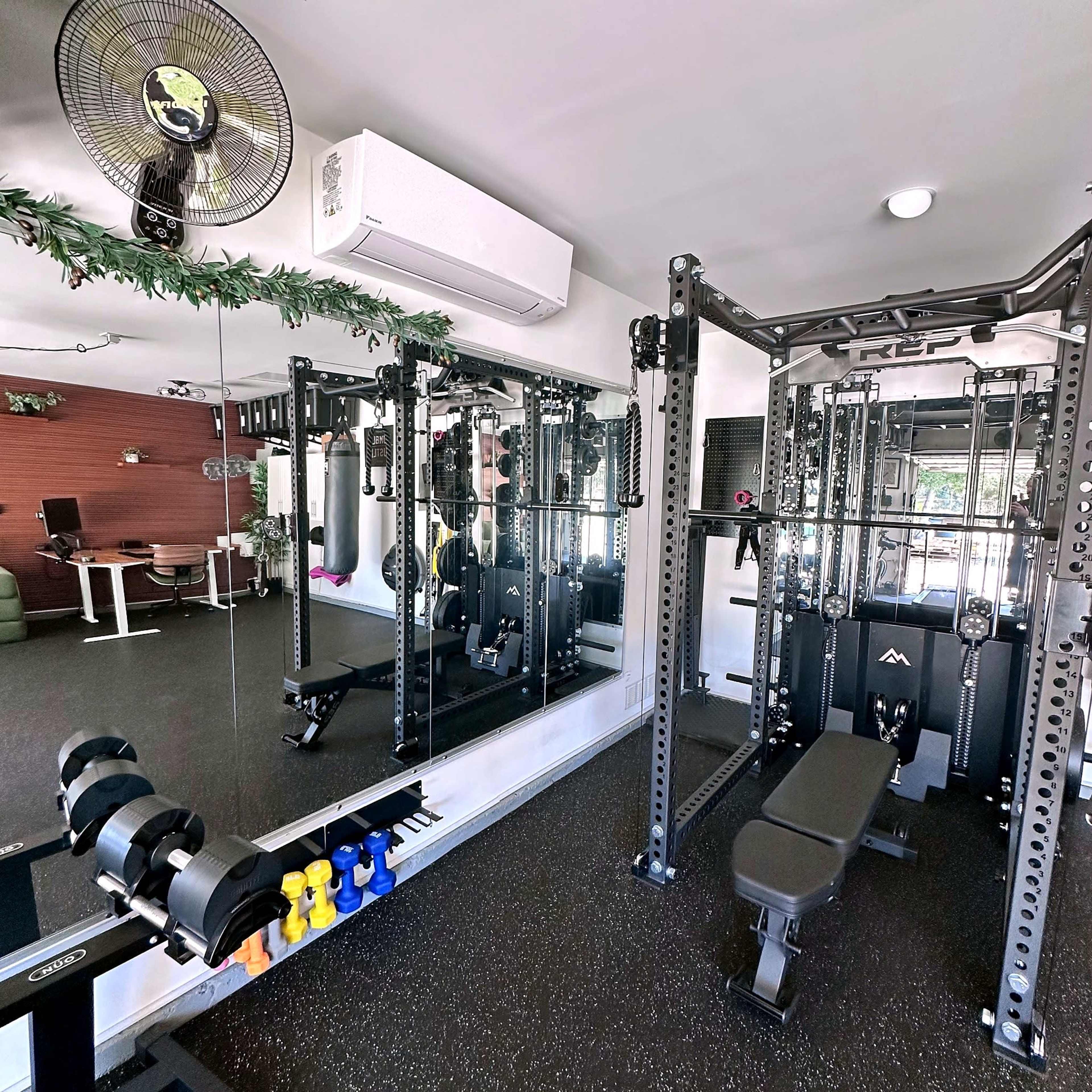 The image shows a home gym with a weightlifting station, free weights, a mirror, and a workout bench.