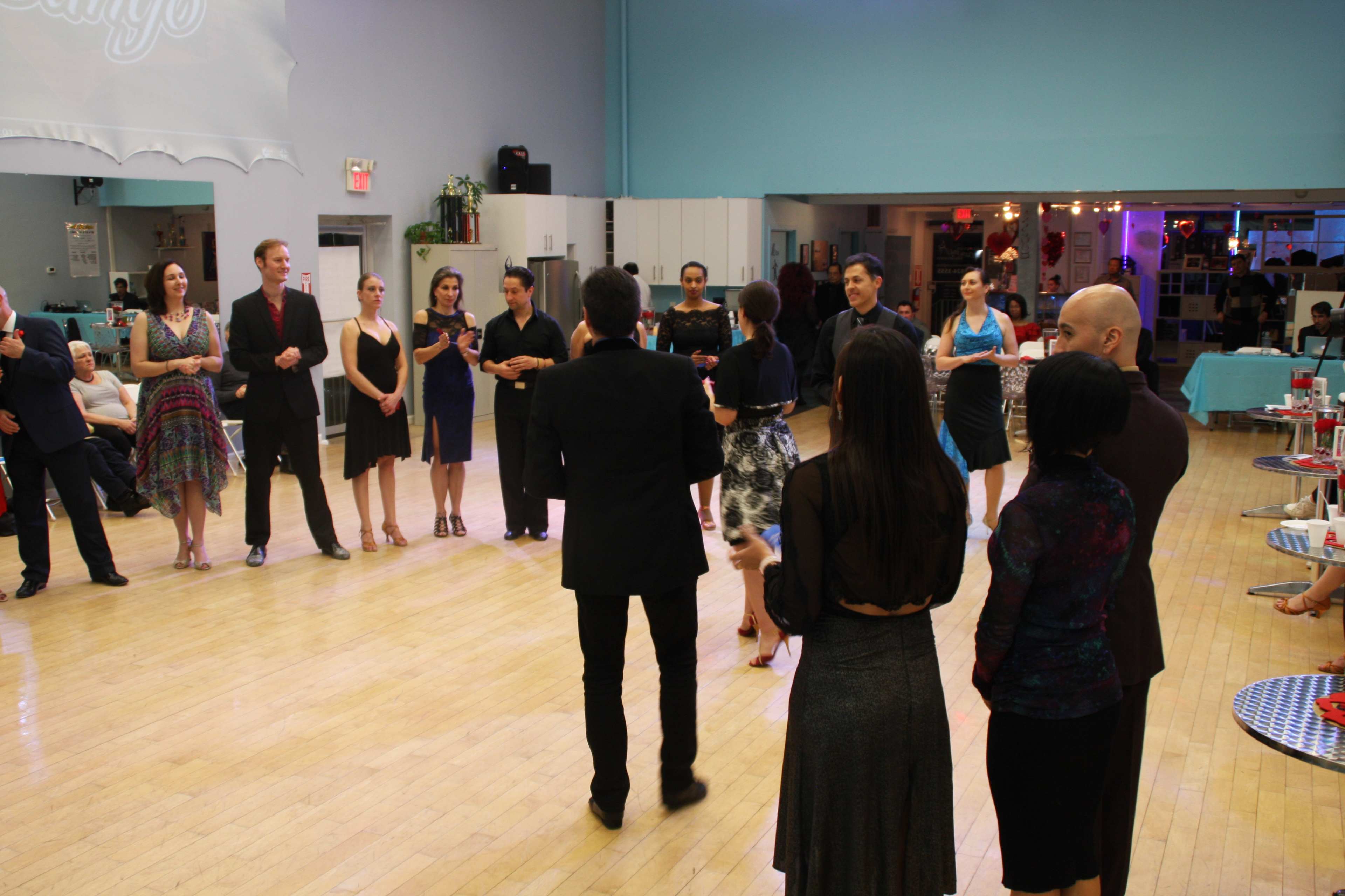 A group of people dressed in formal attire stands in a dance studio, observing a dance performance or competition.