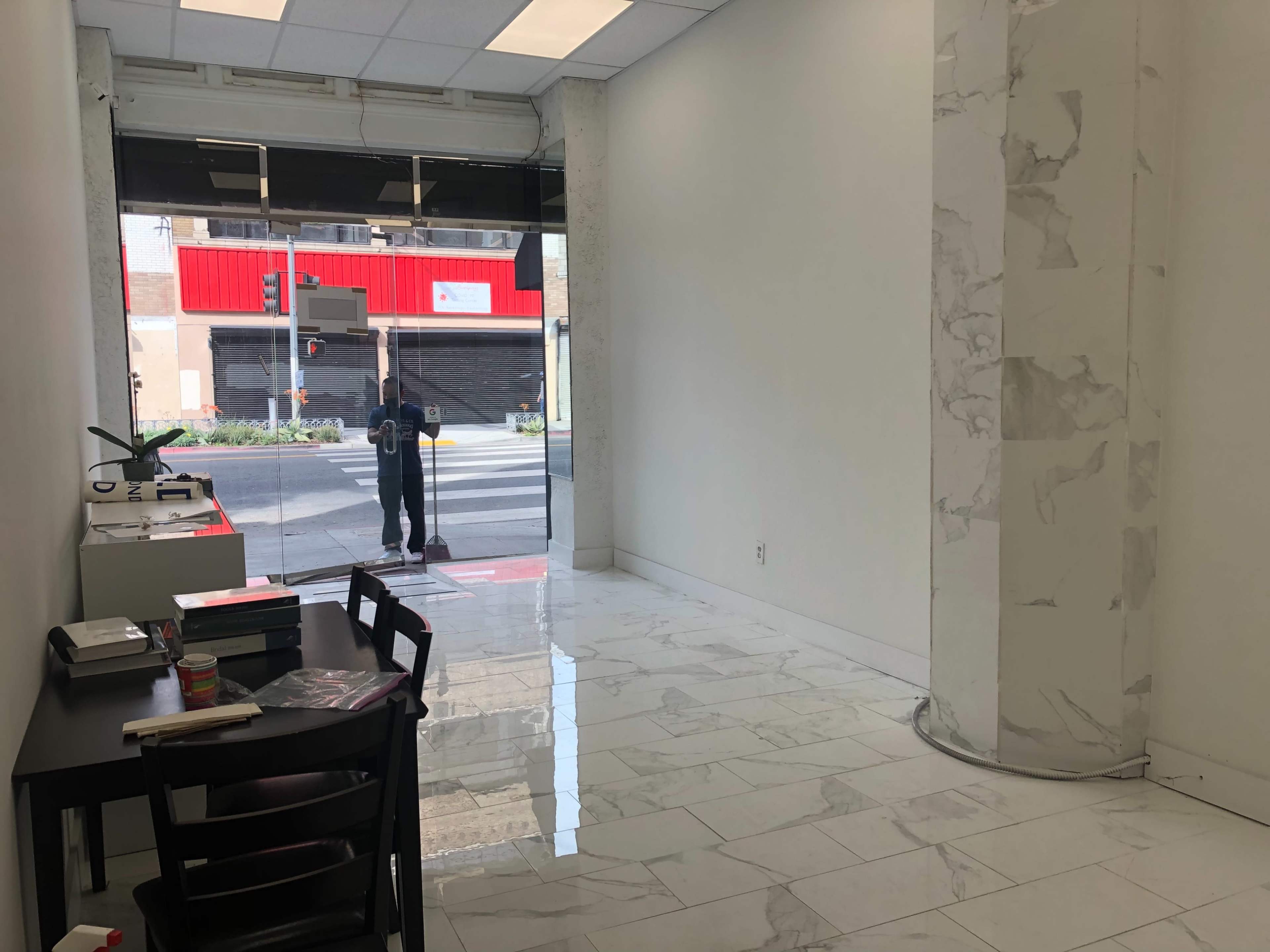 The image shows a bright, minimally furnished interior space with a table and chairs on the left and large windows facing a street outside.