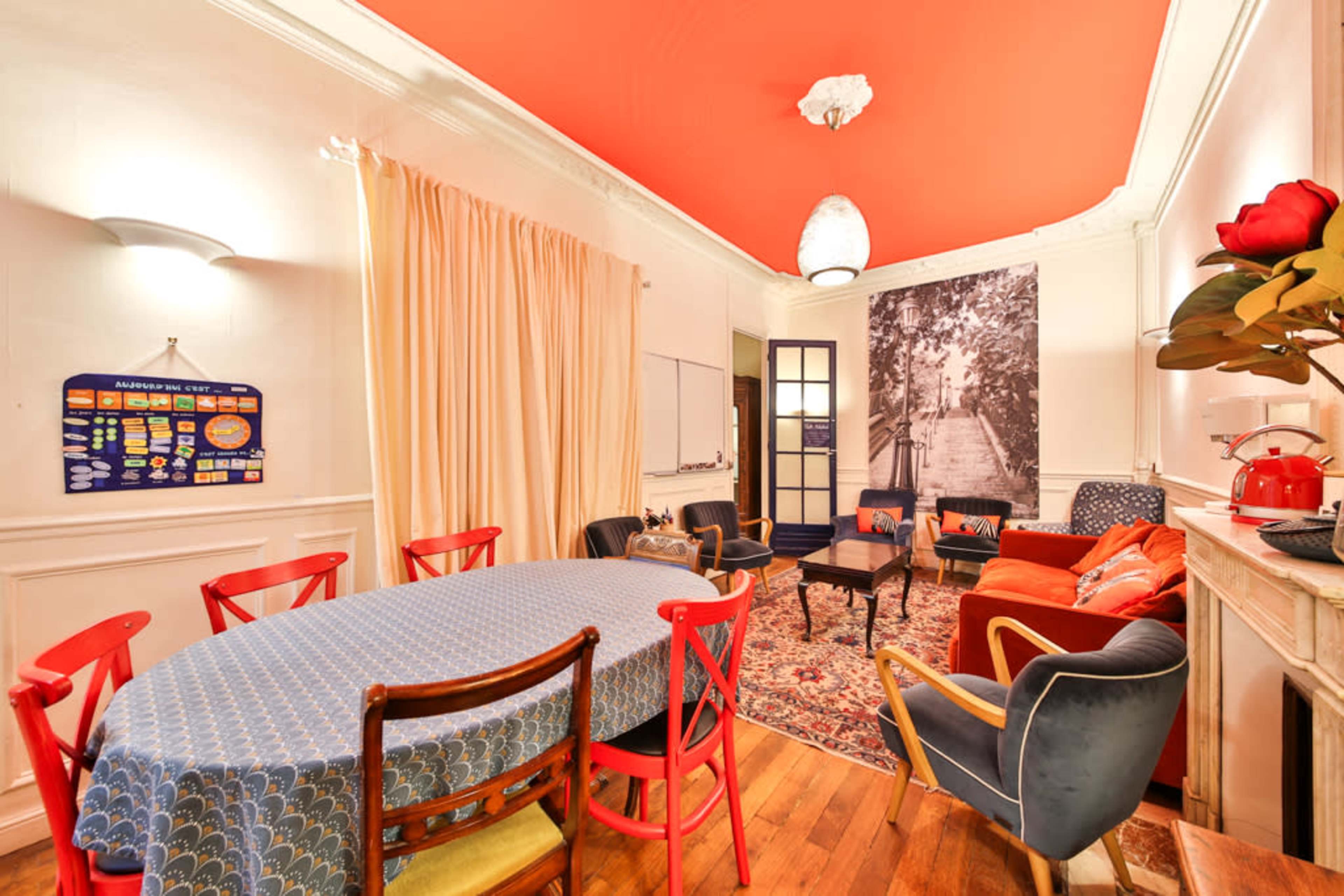 The image shows a brightly decorated room featuring a large table surrounded by chairs, a patterned rug, and various seating areas with a mix of furniture styles.