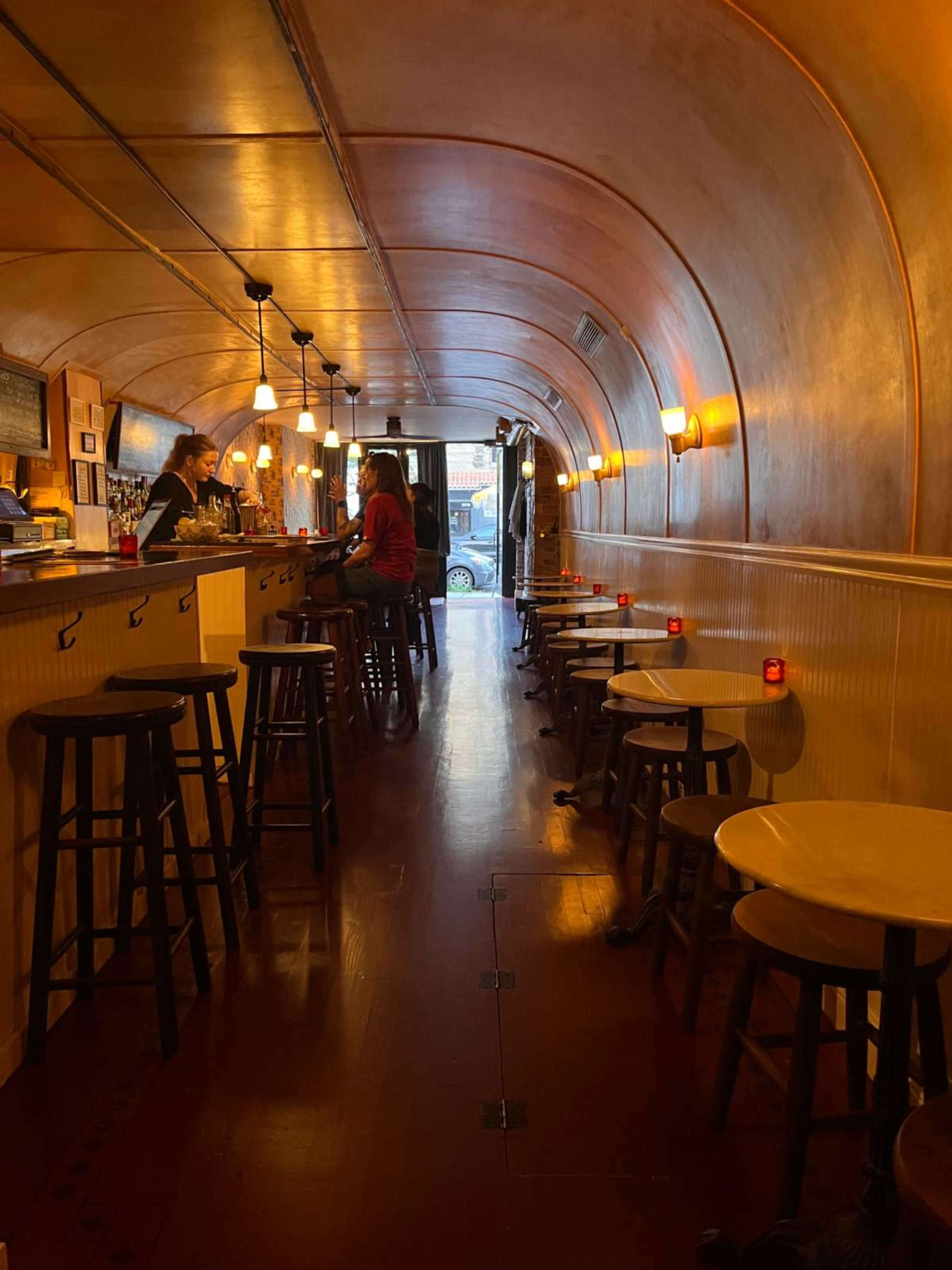 The image shows a narrow, arched interior of a bar with stools arranged along the counter and small tables placed on the floor.