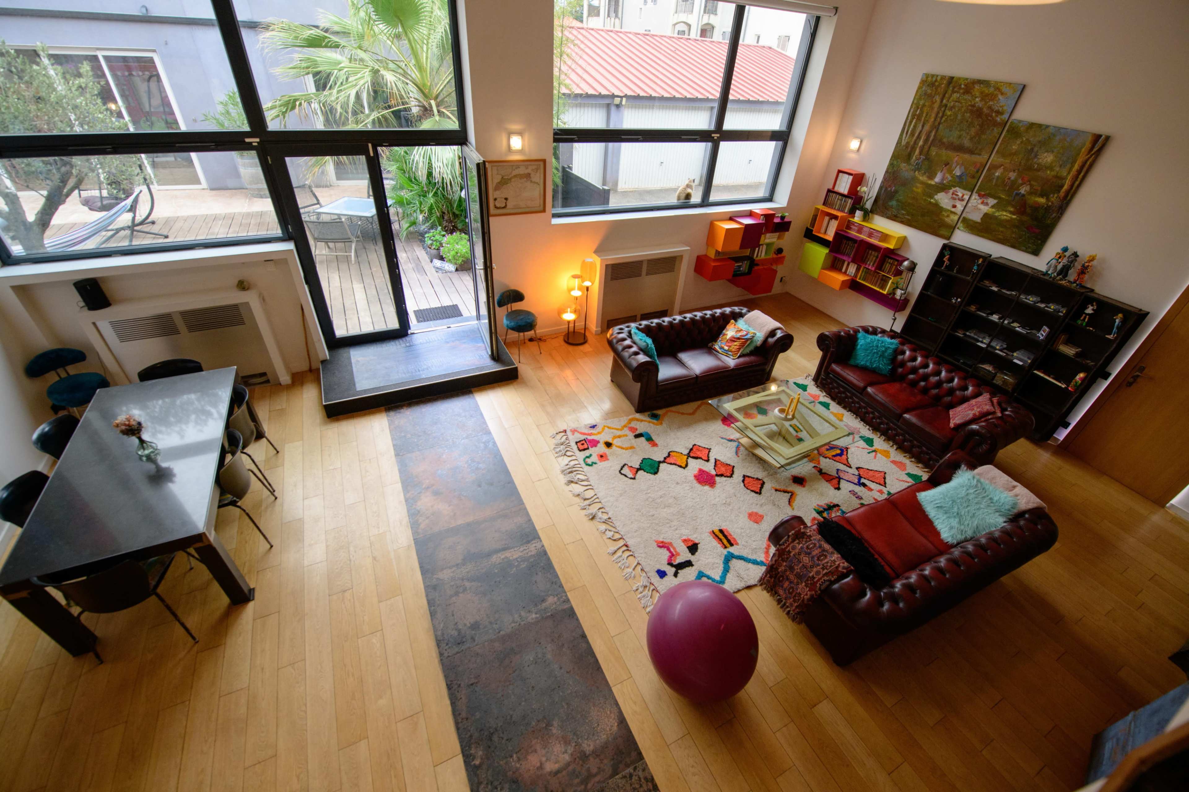 A spacious, modern living area features a large window with a view of a deck, colorful artwork, a patterned rug, and a mix of seating arrangements.