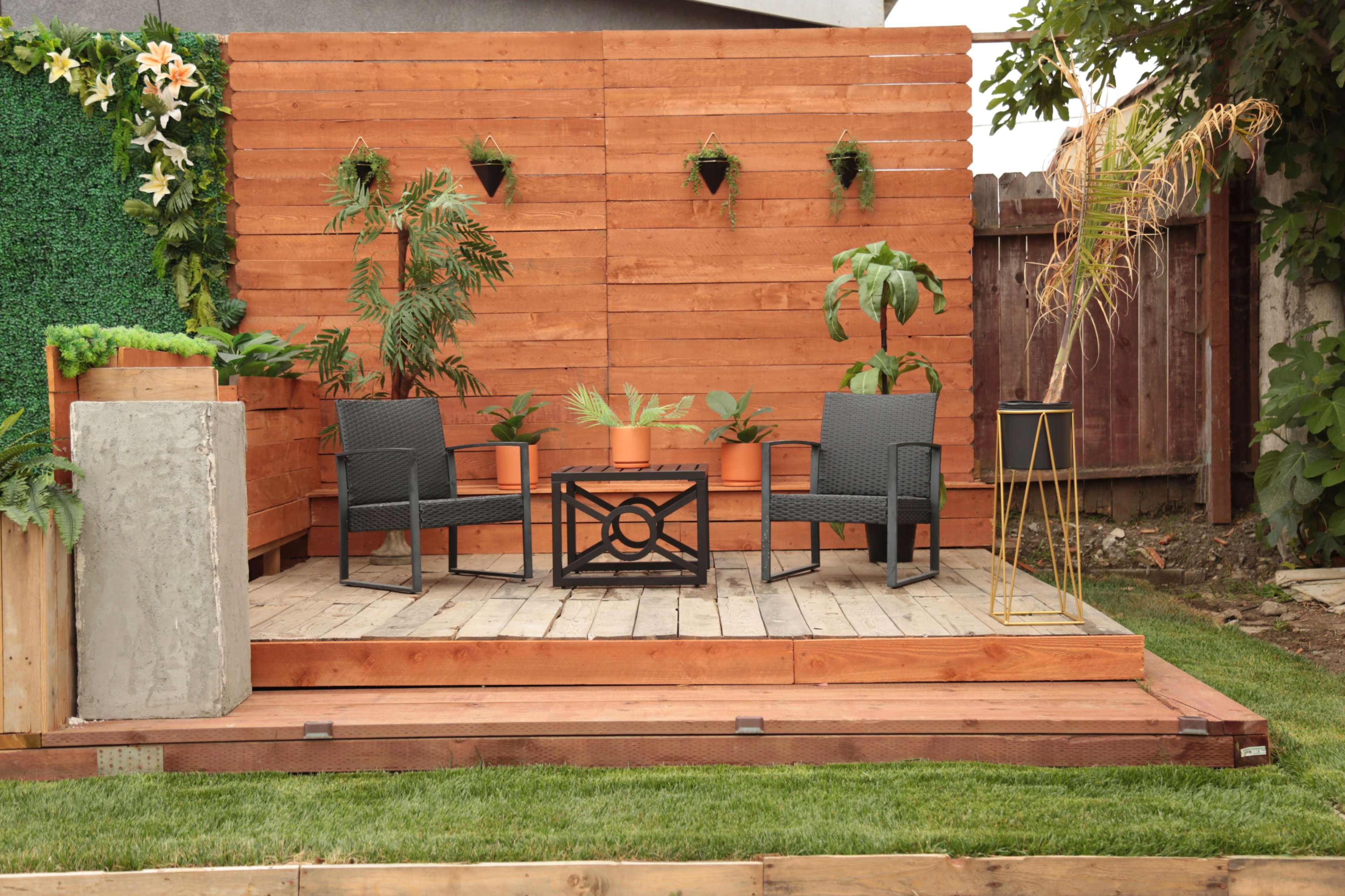 A patio area with two black chairs and a table surrounded by wooden panels, potted plants, and a grassy lawn.