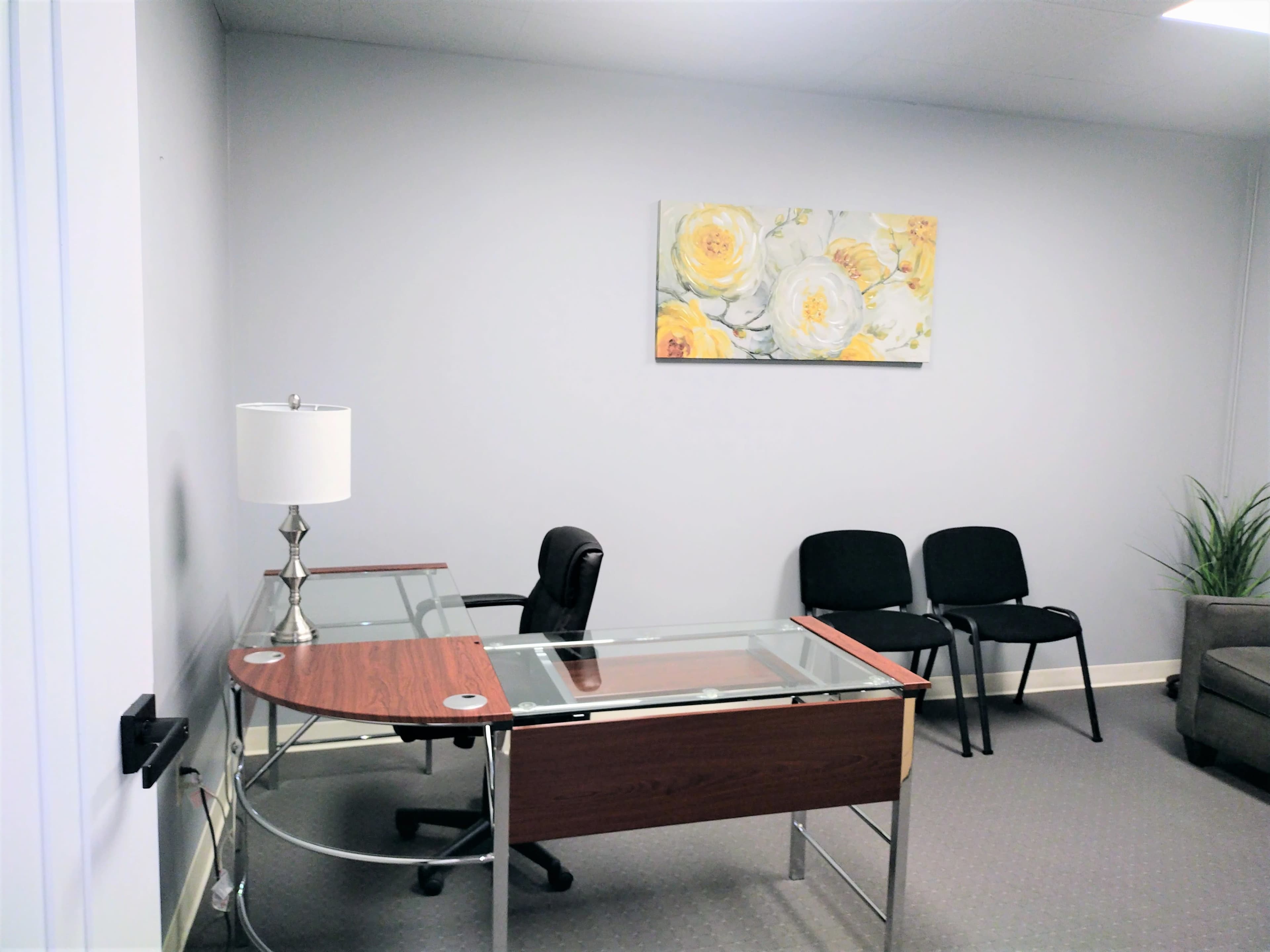 A small office space featuring a glass-topped desk, a black office chair, two black chairs, and a floral painting on the wall.