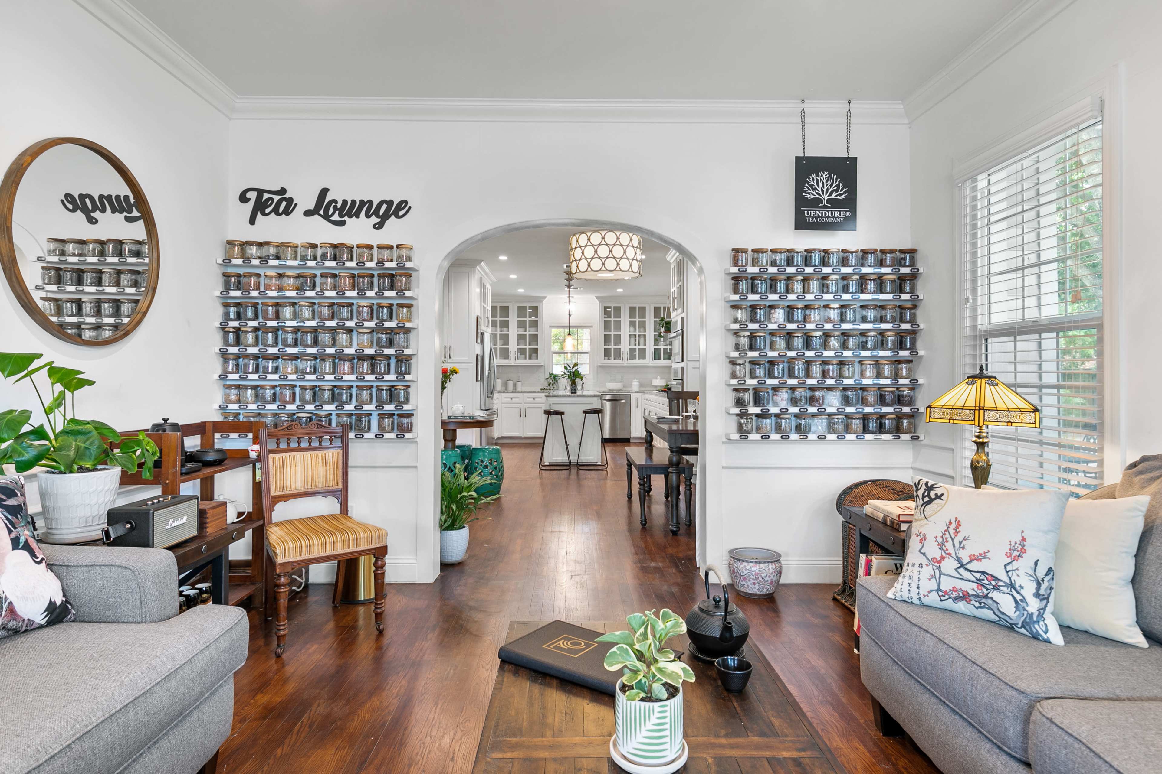A cozy living area leading into a bright kitchen, featuring a tea lounge sign, a wall of jars, and comfortable seating.
