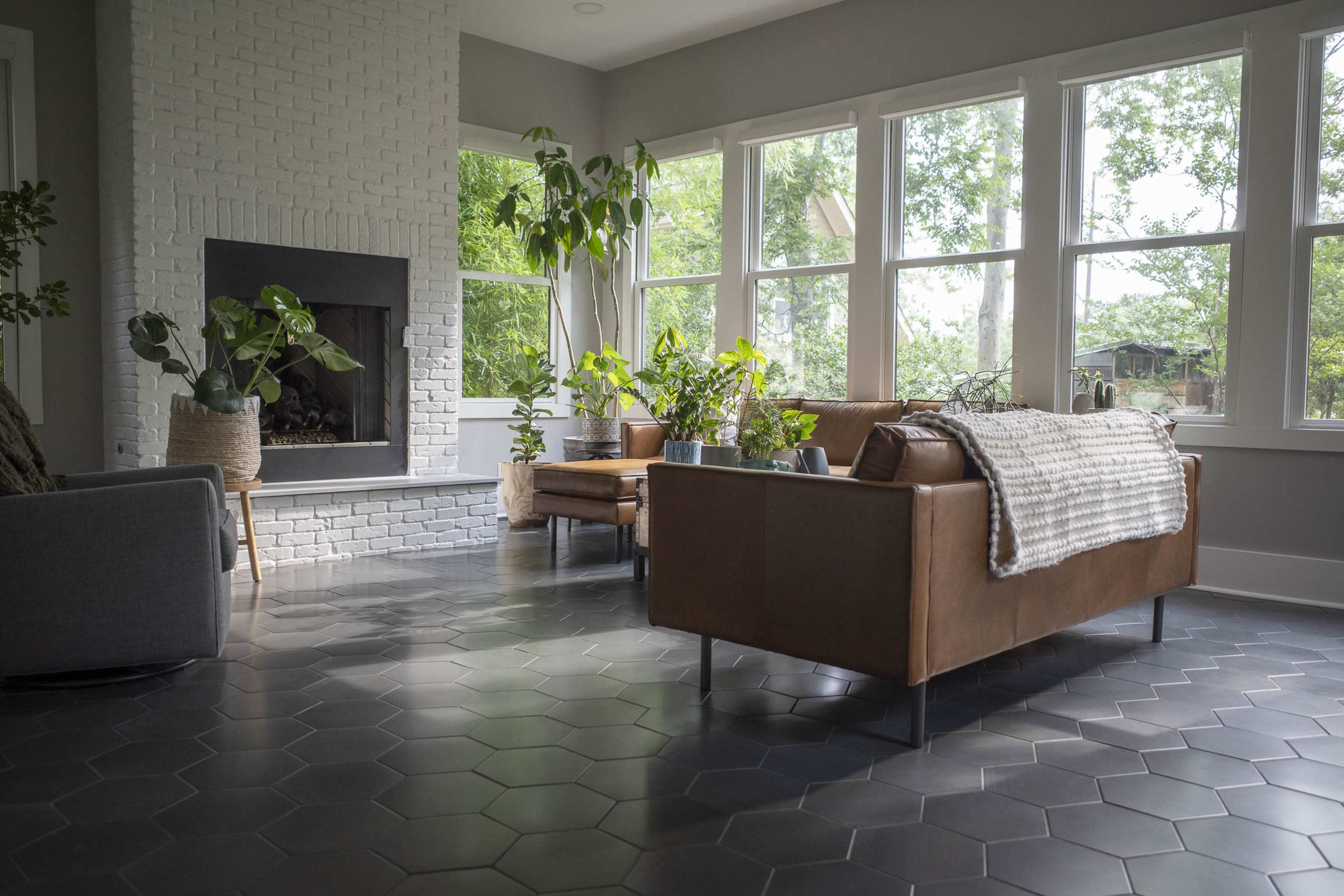 A modern living room with a large window, a brick fireplace, and plants arranged around leather and fabric furniture on hexagonal tile flooring.