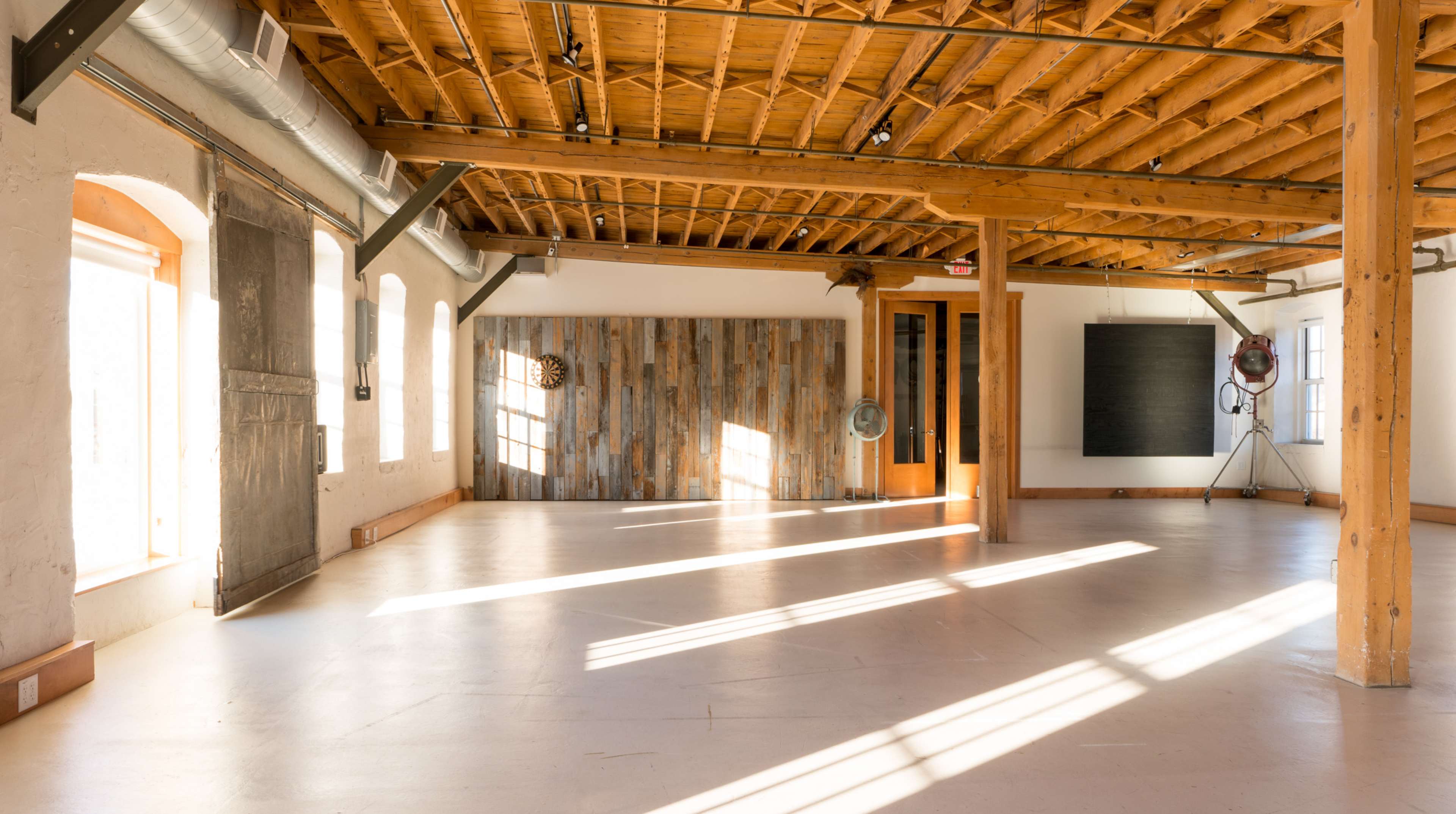 The image shows a spacious, well-lit studio with wooden beams and a polished concrete floor, featuring large windows and a wooden accent wall.