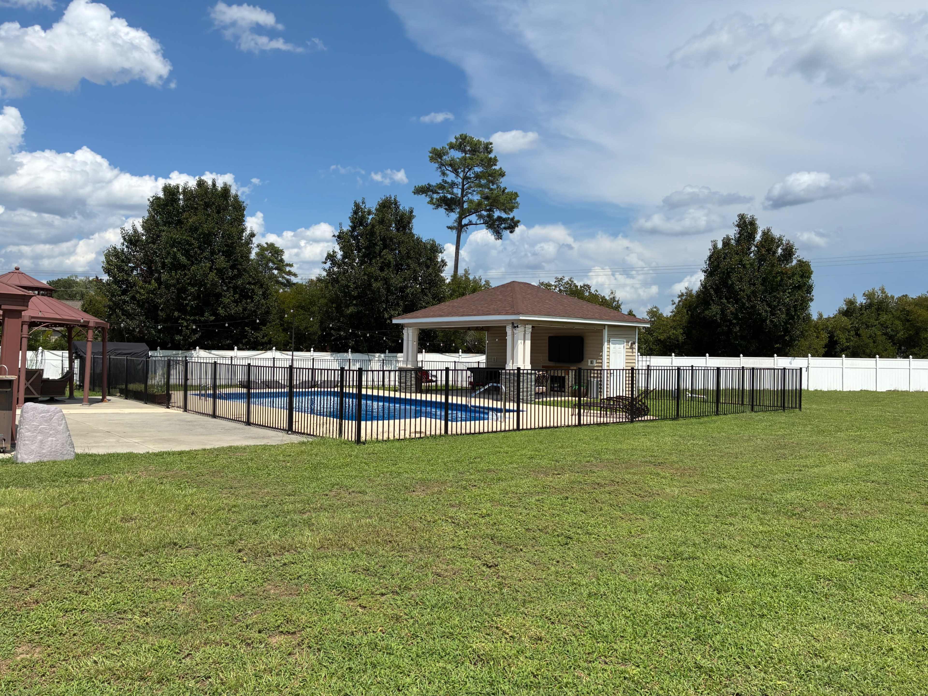 The image shows a fenced-in backyard with a swimming pool, a covered patio, and a well-maintained lawn under a blue sky with scattered clouds.