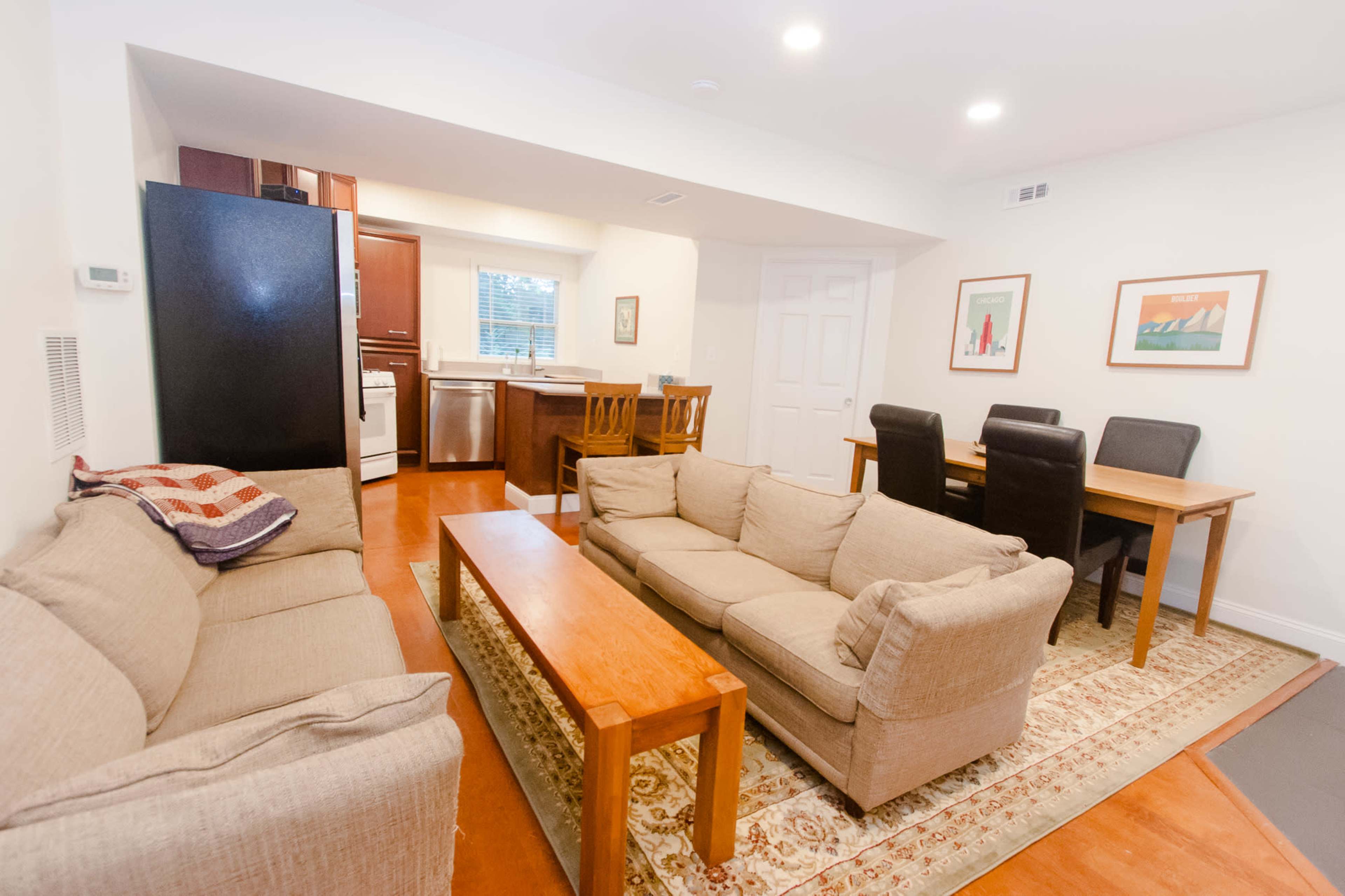 A living space featuring a seating area with two couches, a wooden coffee table, a dining table with chairs, and a kitchenette with appliances.