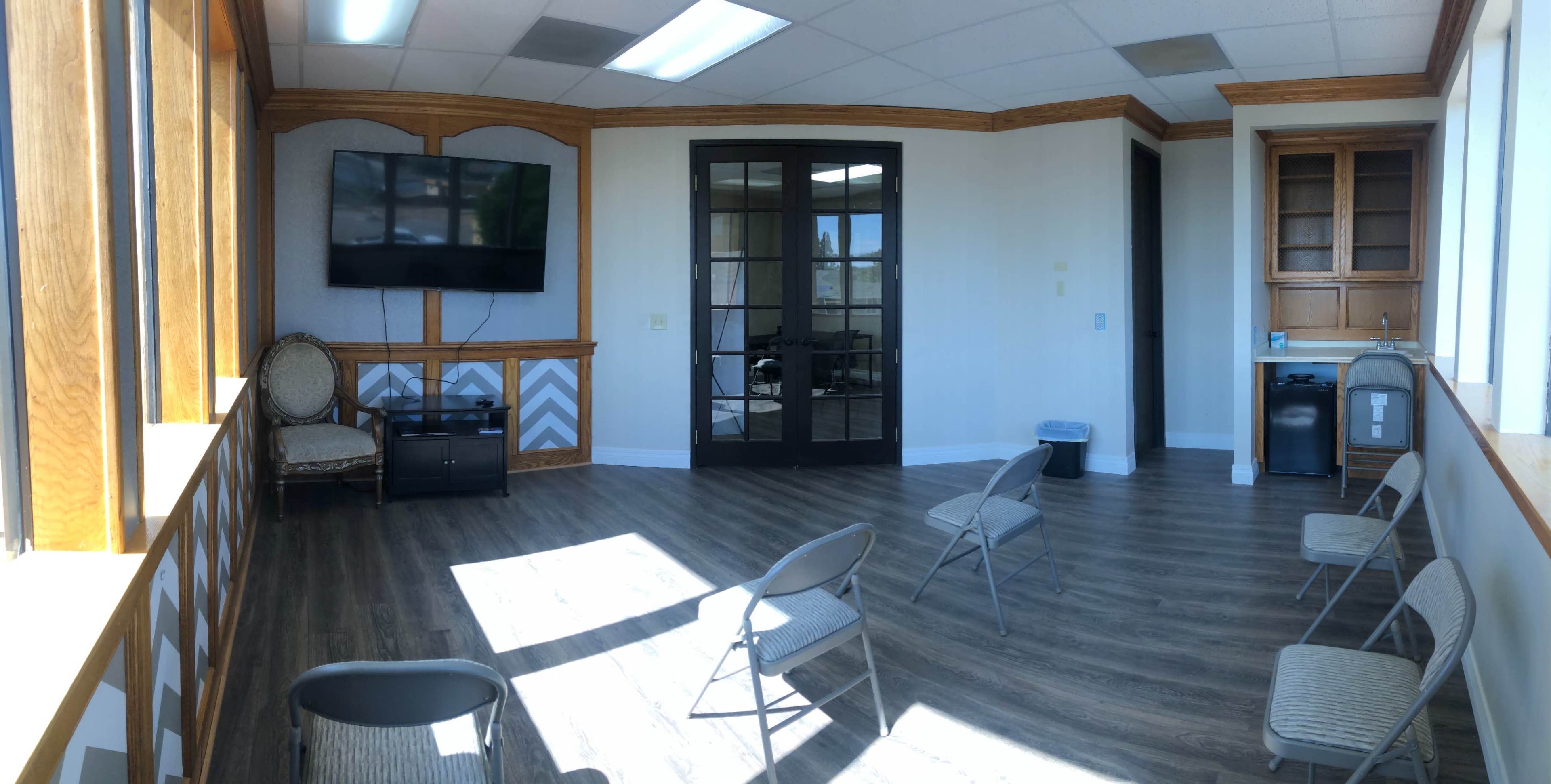 A waiting room features several metal chairs arranged in a circular formation, with a large television mounted on the wall and a glass door leading to another room.