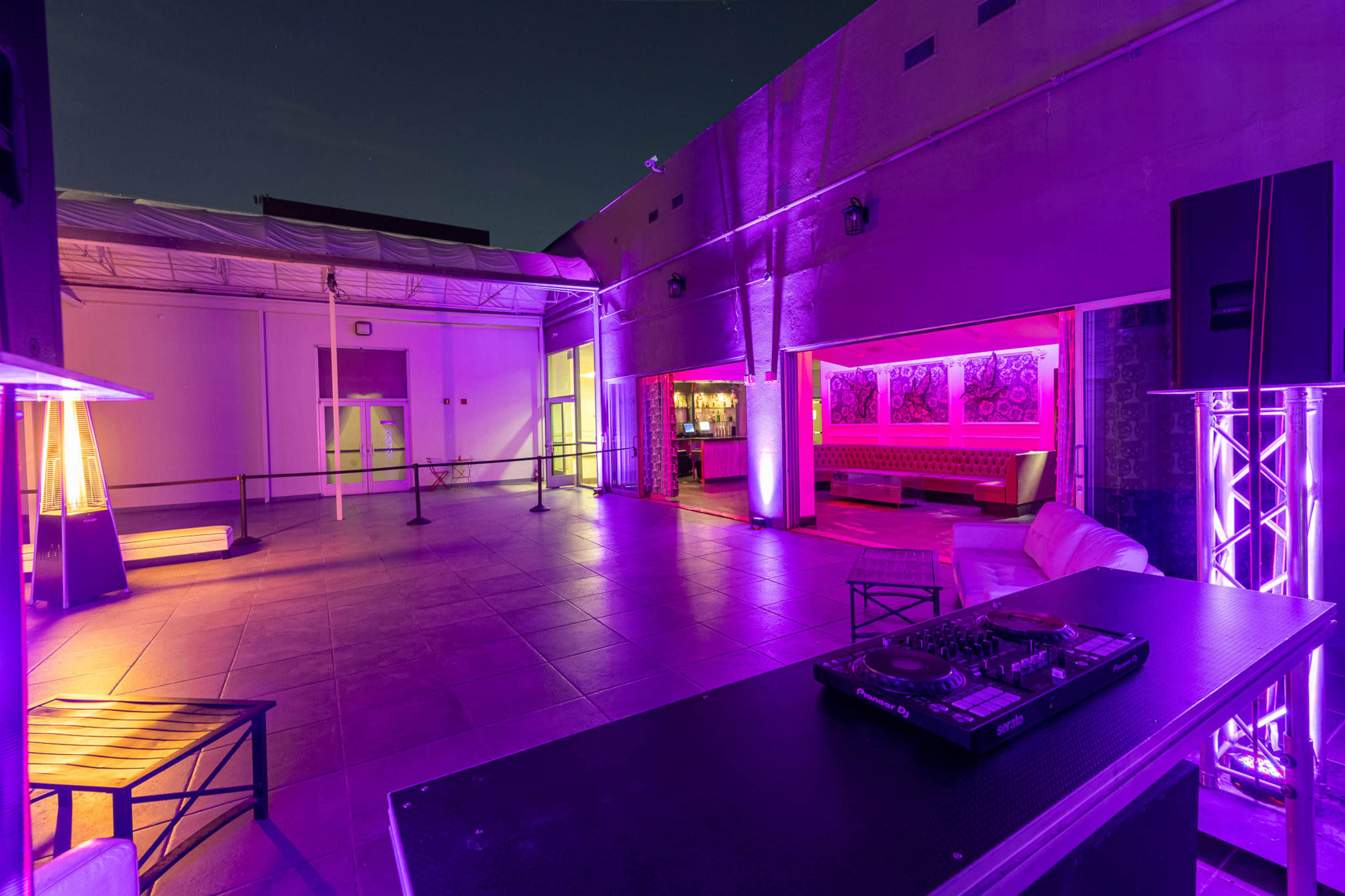 The image shows a modern outdoor lounge area with a DJ setup, illuminated by purple lights, and featuring a bar in the background.