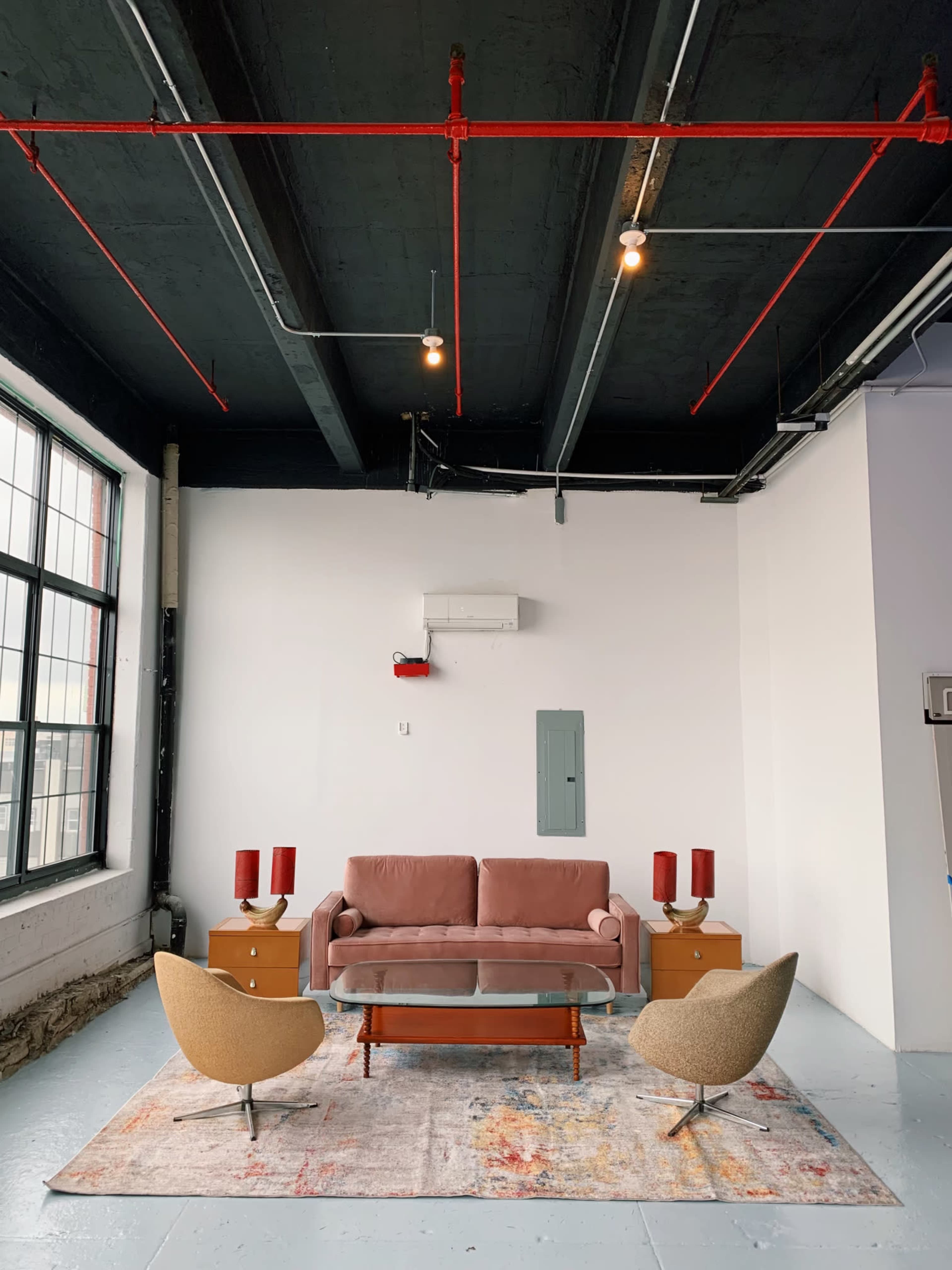 A minimalist living room with a pink sofa, two armchairs, a coffee table, and red table lamps, set against a white wall and a ceiling with exposed pipes and lights.