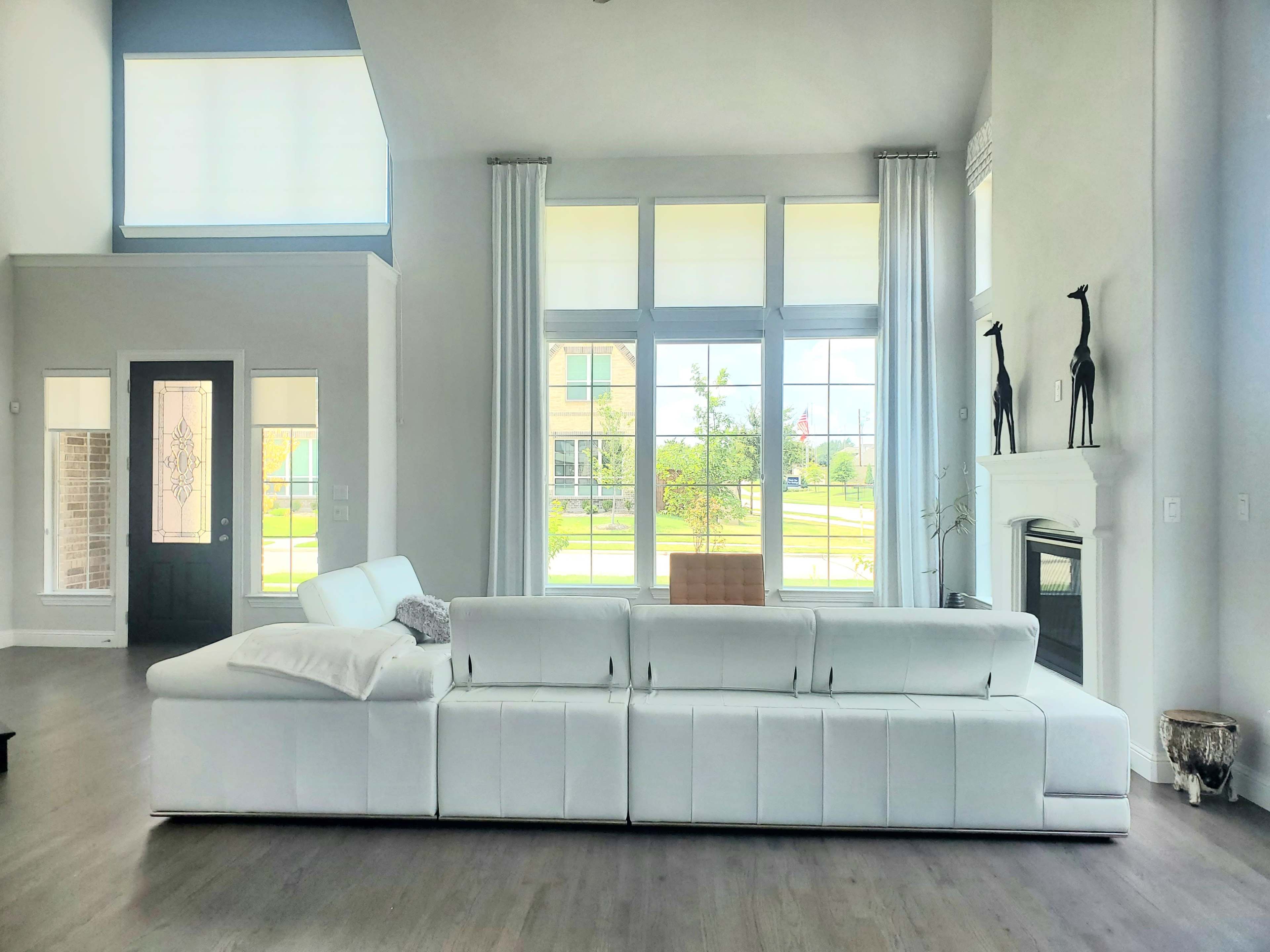 A modern living room features a large white sectional sofa facing tall windows with natural light streaming in.