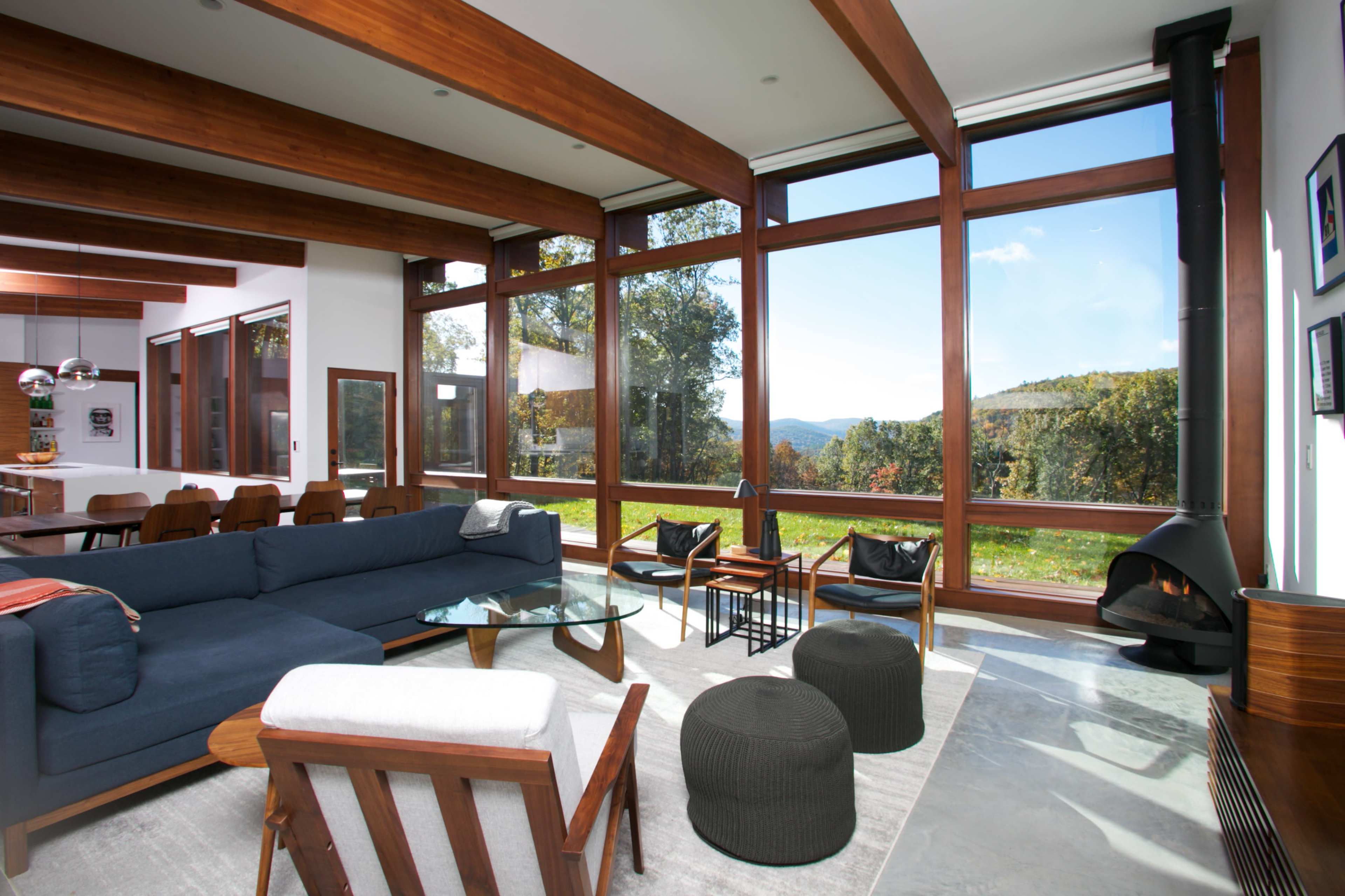 The scene features a modern living room with large windows offering a view of the landscape, furnished with a blue sofa, glass coffee table, and black accent pieces.