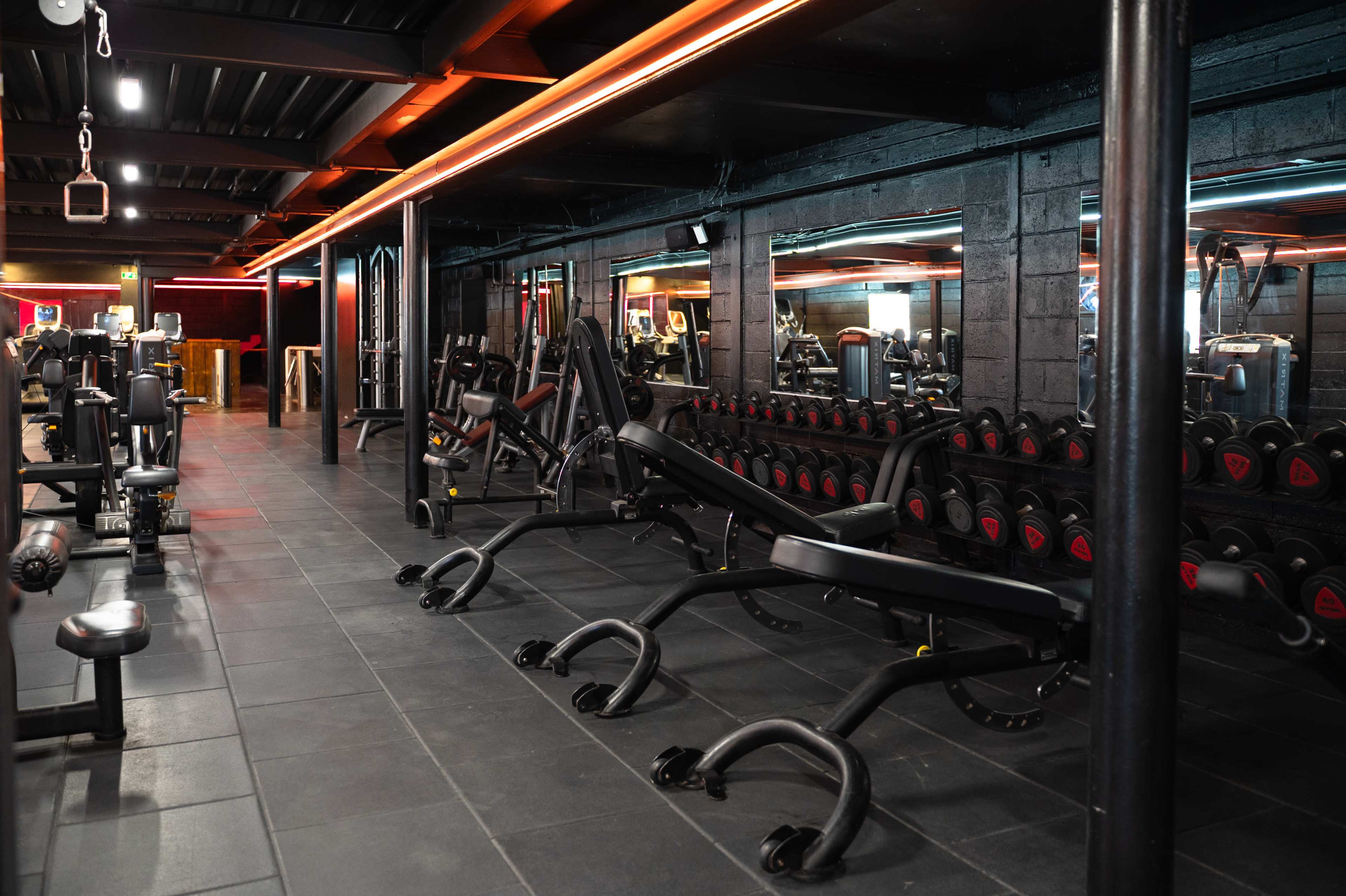 The image shows a well-equipped gym interior featuring various exercise machines and free weights arranged neatly along the walls.