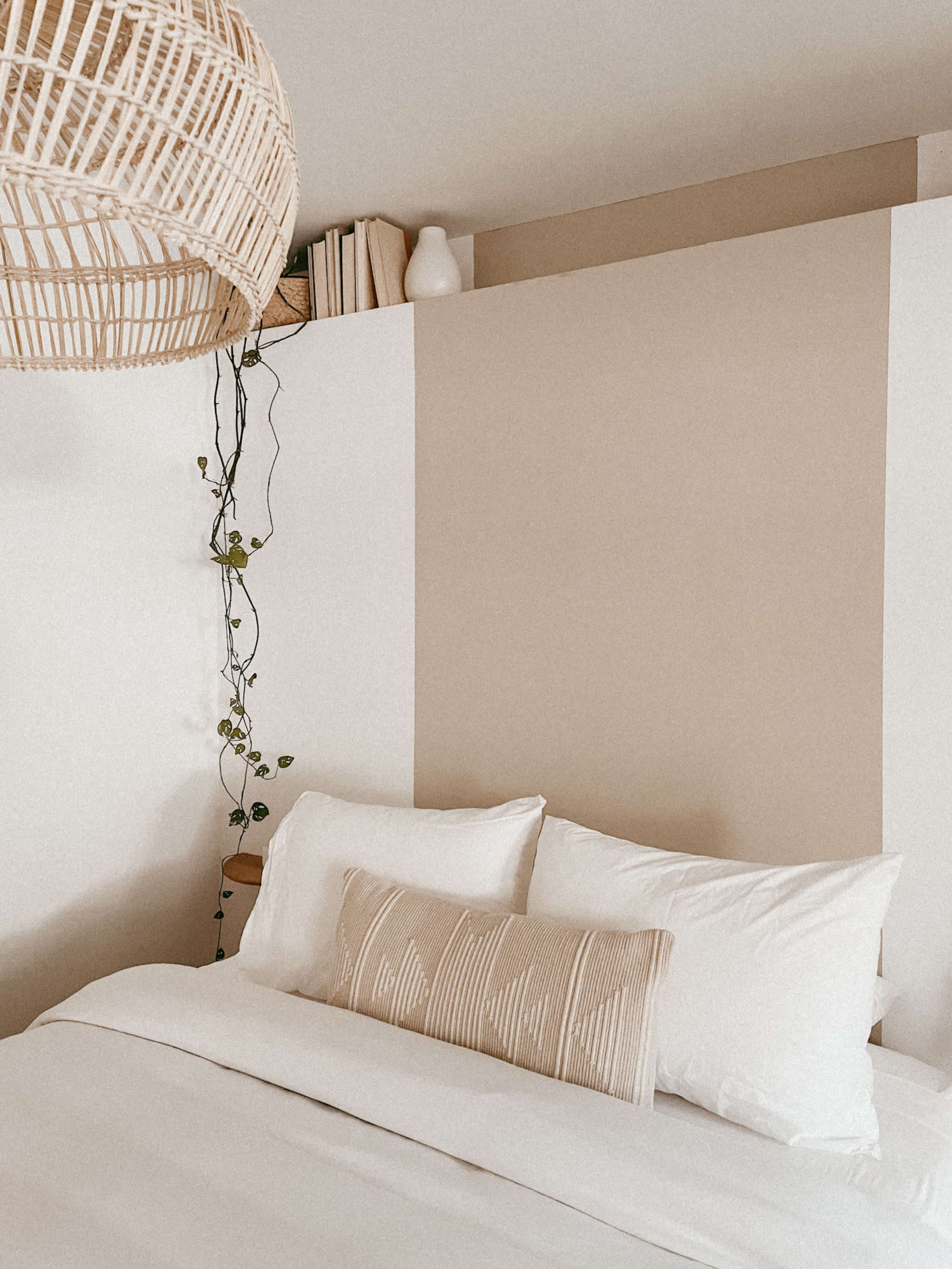 A cozy bedroom with a bed featuring white linens and a decorative pillow, surrounded by neutral-colored walls and a hanging woven light fixture.