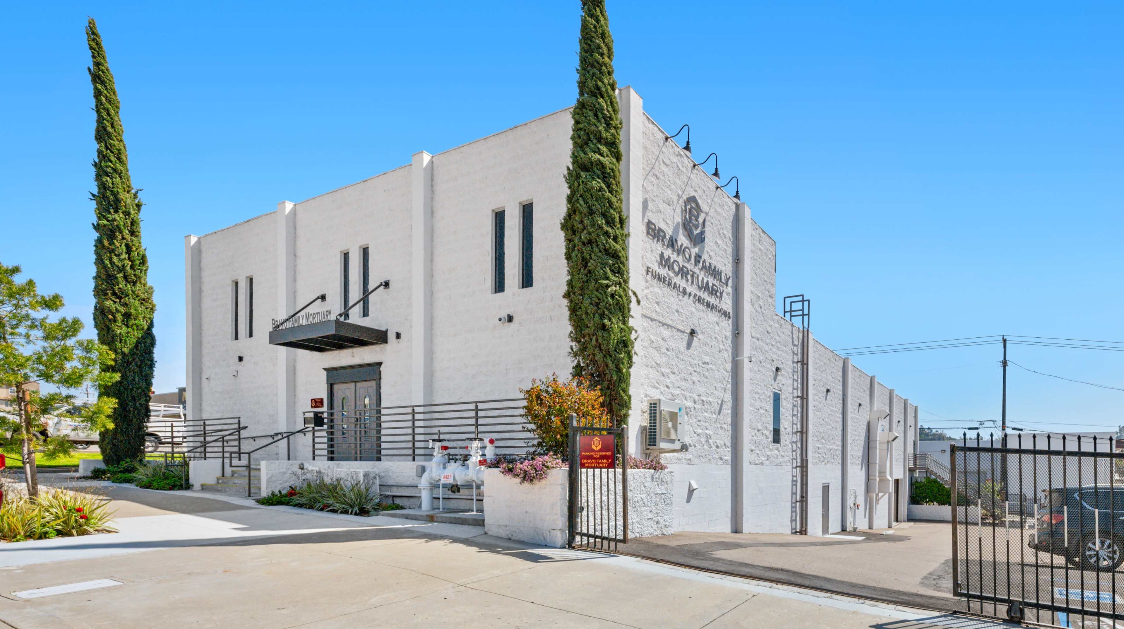 The image shows a white, modern building with tall cypress trees on either side and a gated entrance.