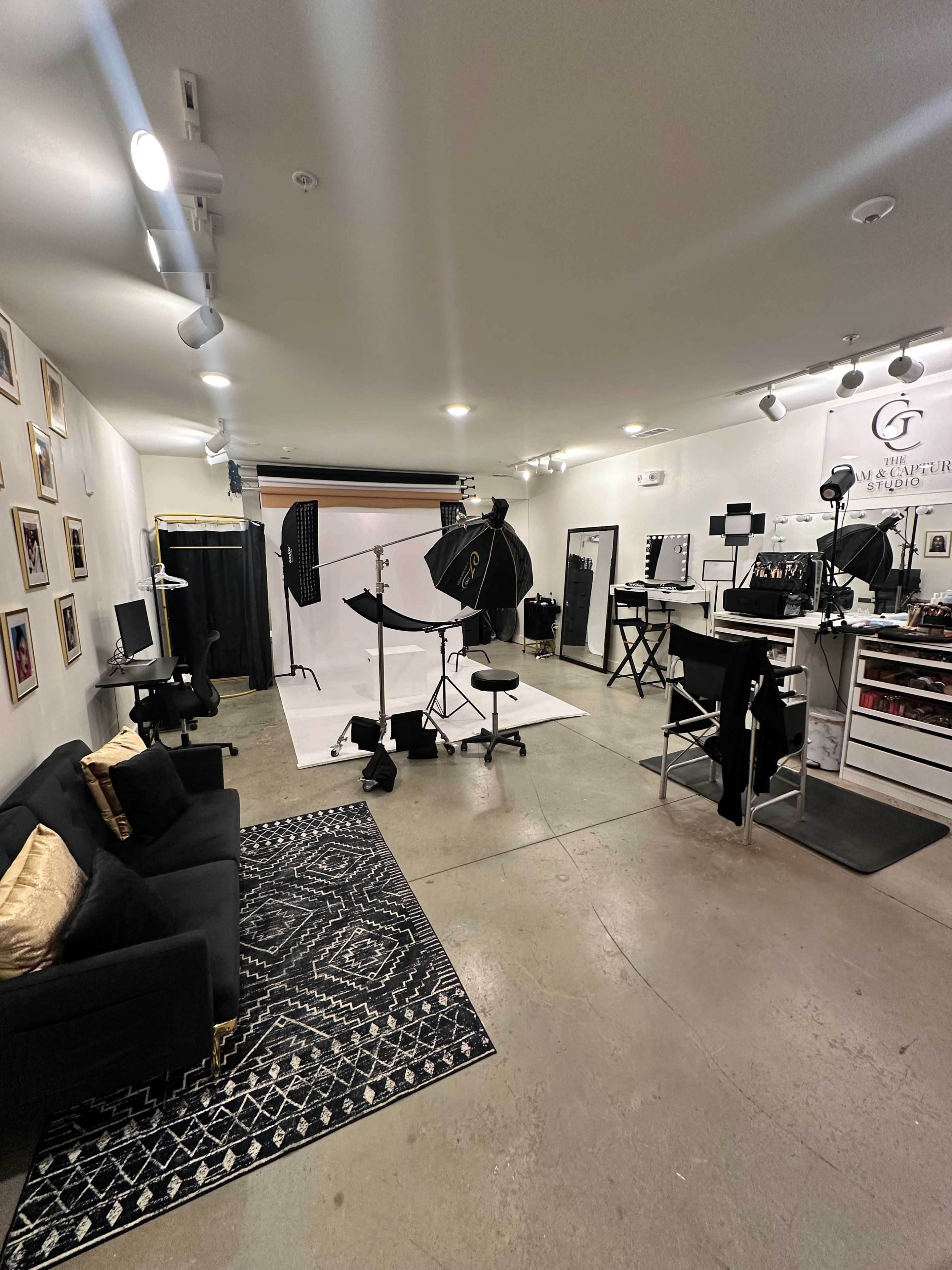 The image shows a photography studio featuring a white backdrop, lighting equipment, makeup stations, and a black sofa on a patterned rug.