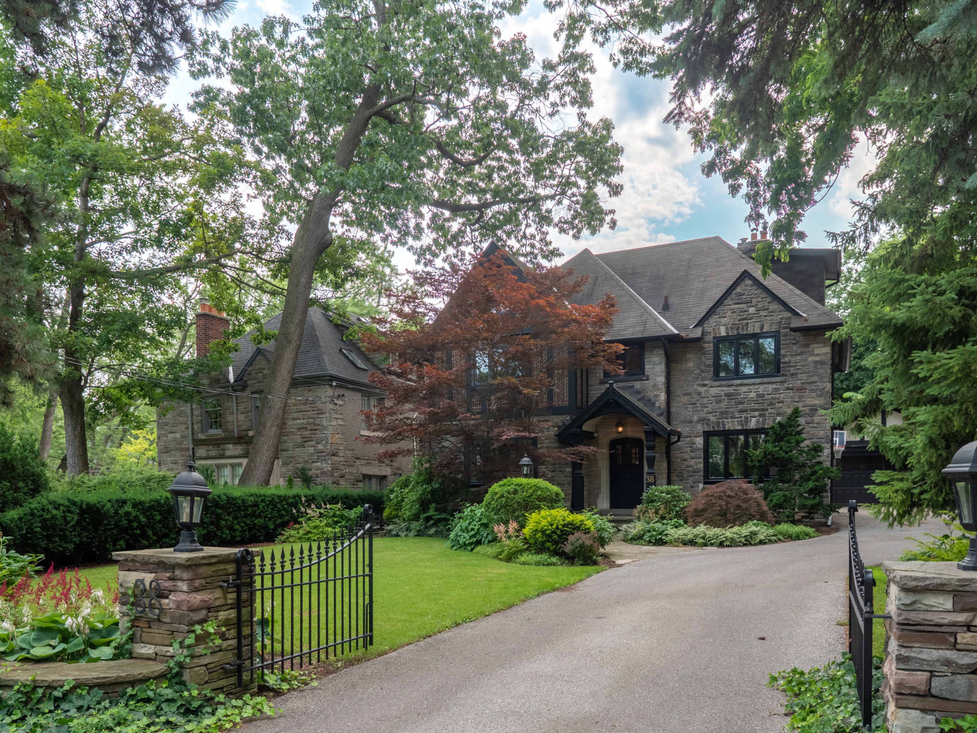 A large stone house with a triangular roof is surrounded by lush greenery and a well-maintained garden, featuring a winding driveway and gated entrance.