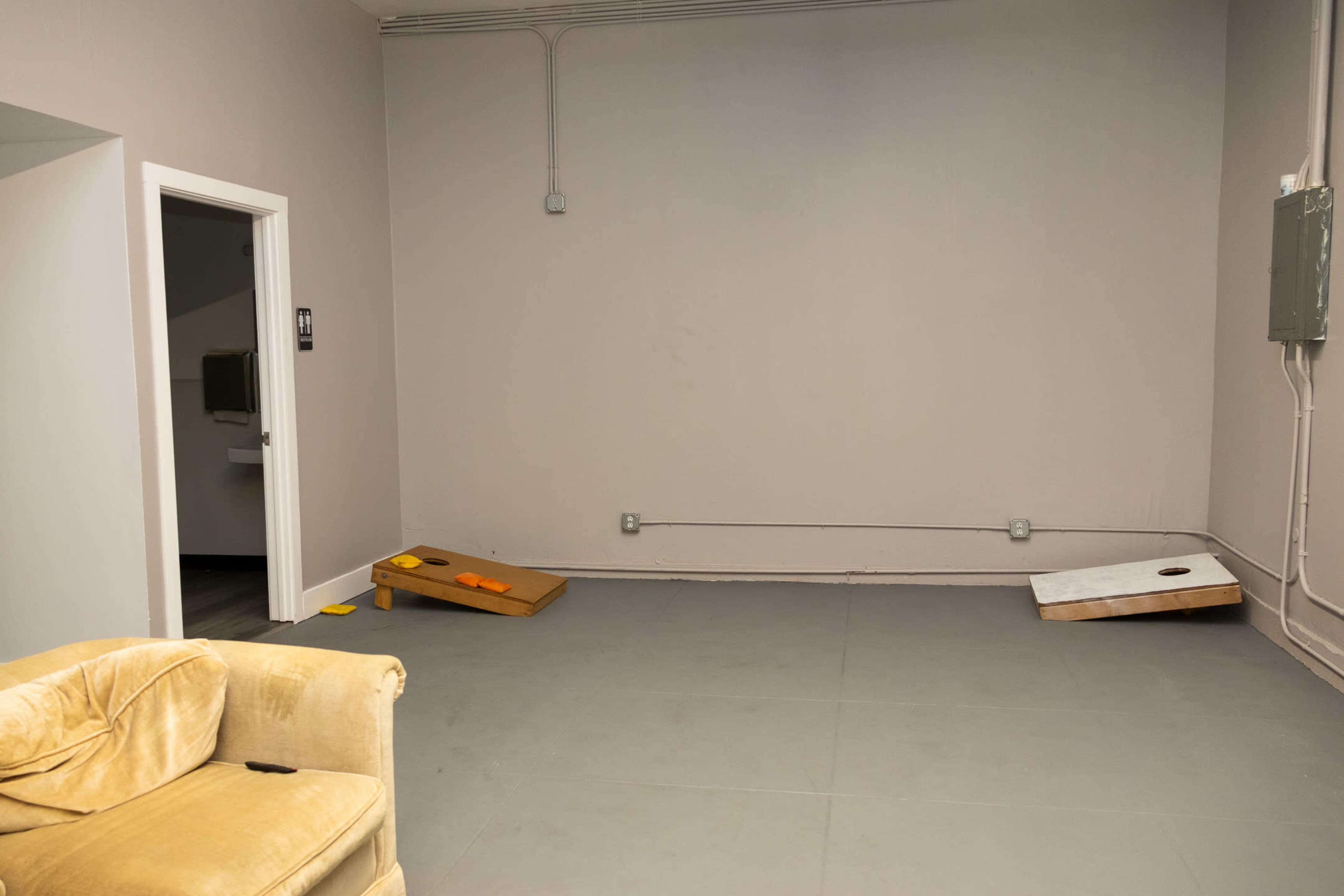 The image shows a vacant room with a light-colored floor, a yellow couch, and two wooden cornhole boards positioned at an angle against the wall.
