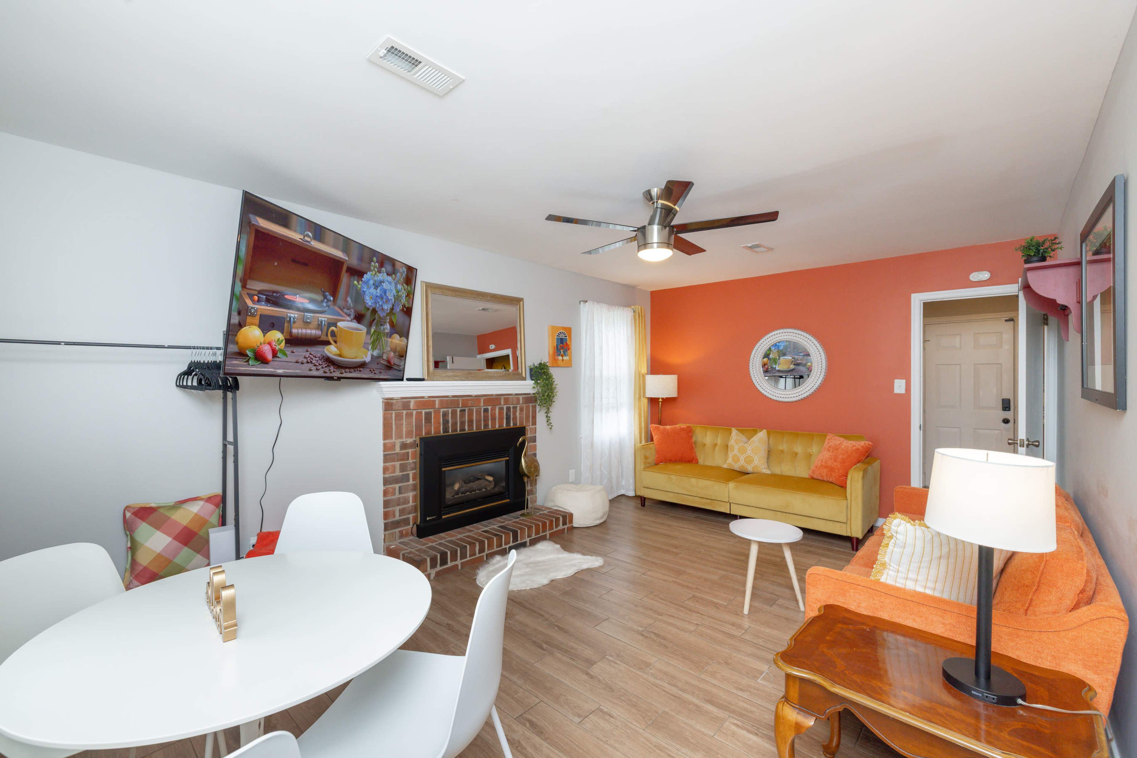 The image depicts a cozy living room featuring an orange wall, a fireplace with a TV mounted above it, a yellow sofa, an orange chair, and a round dining table with white chairs.