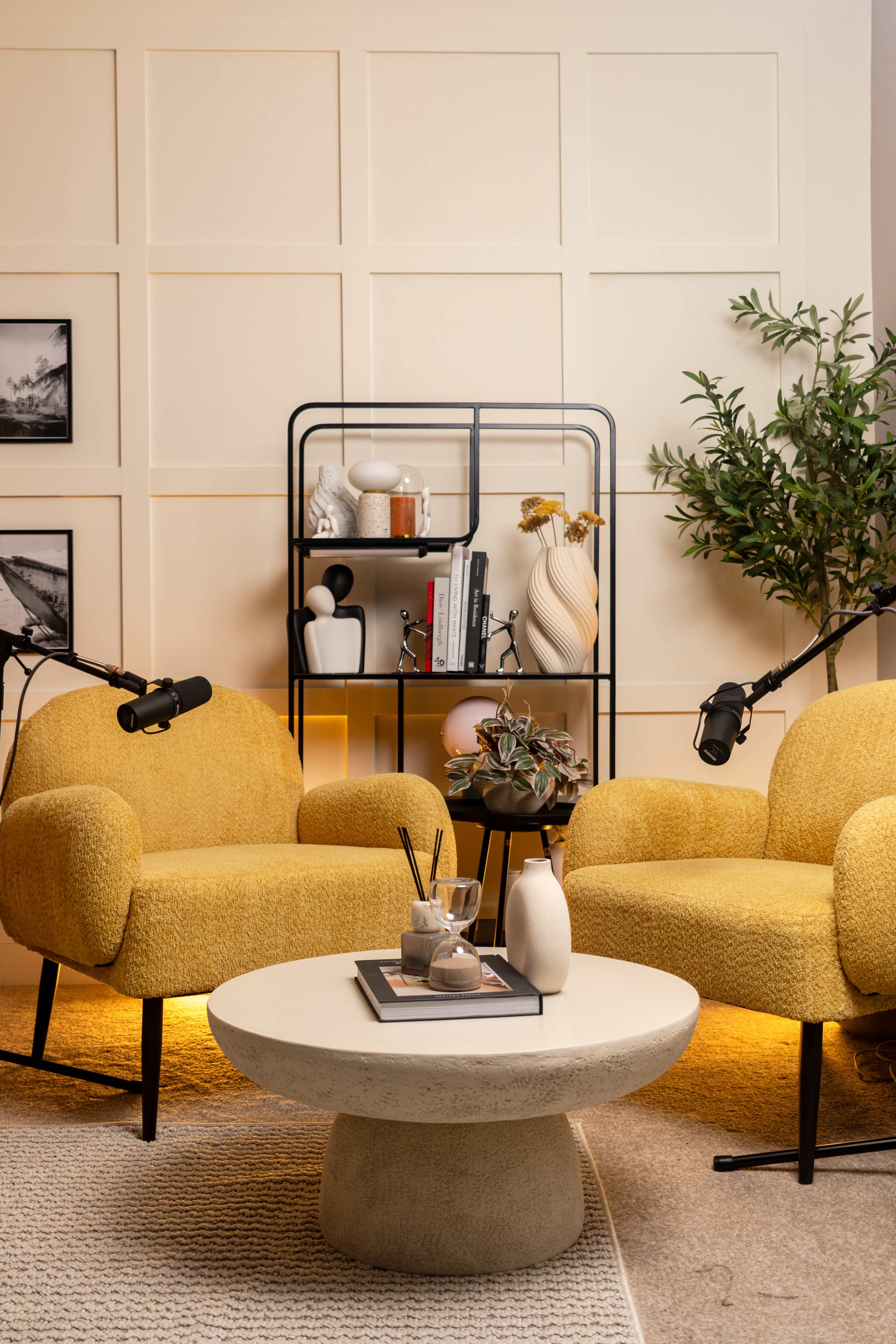 The image features two yellow armchairs facing a round concrete coffee table, framed by a decorative shelving unit with various objects, and a potted plant nearby.