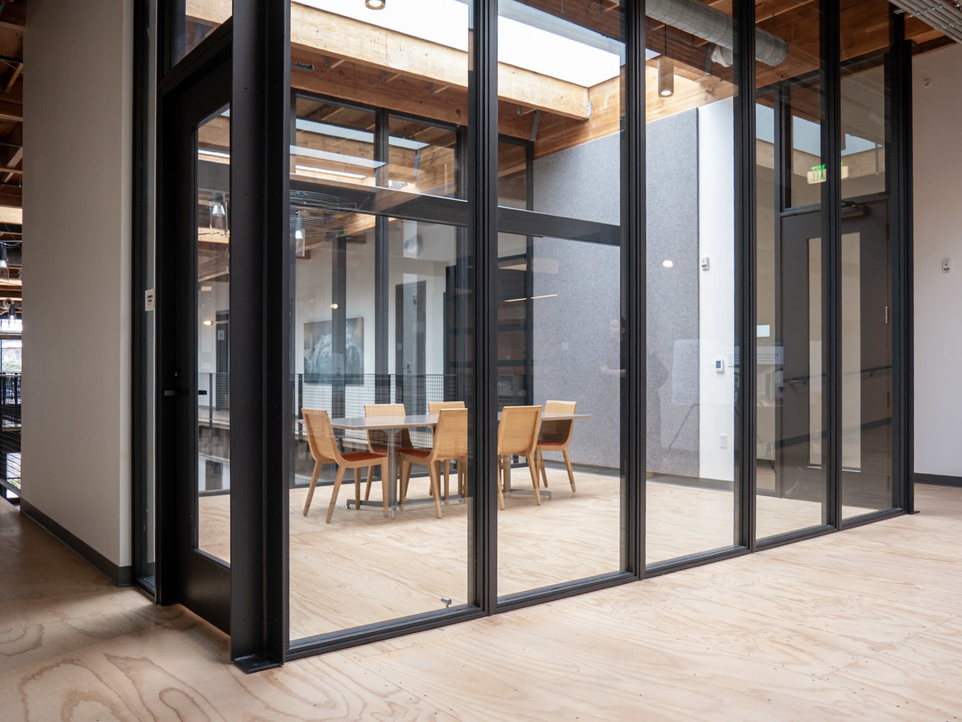 The image shows a meeting room with a large wooden table surrounded by chairs, enclosed by glass walls in an office space.
